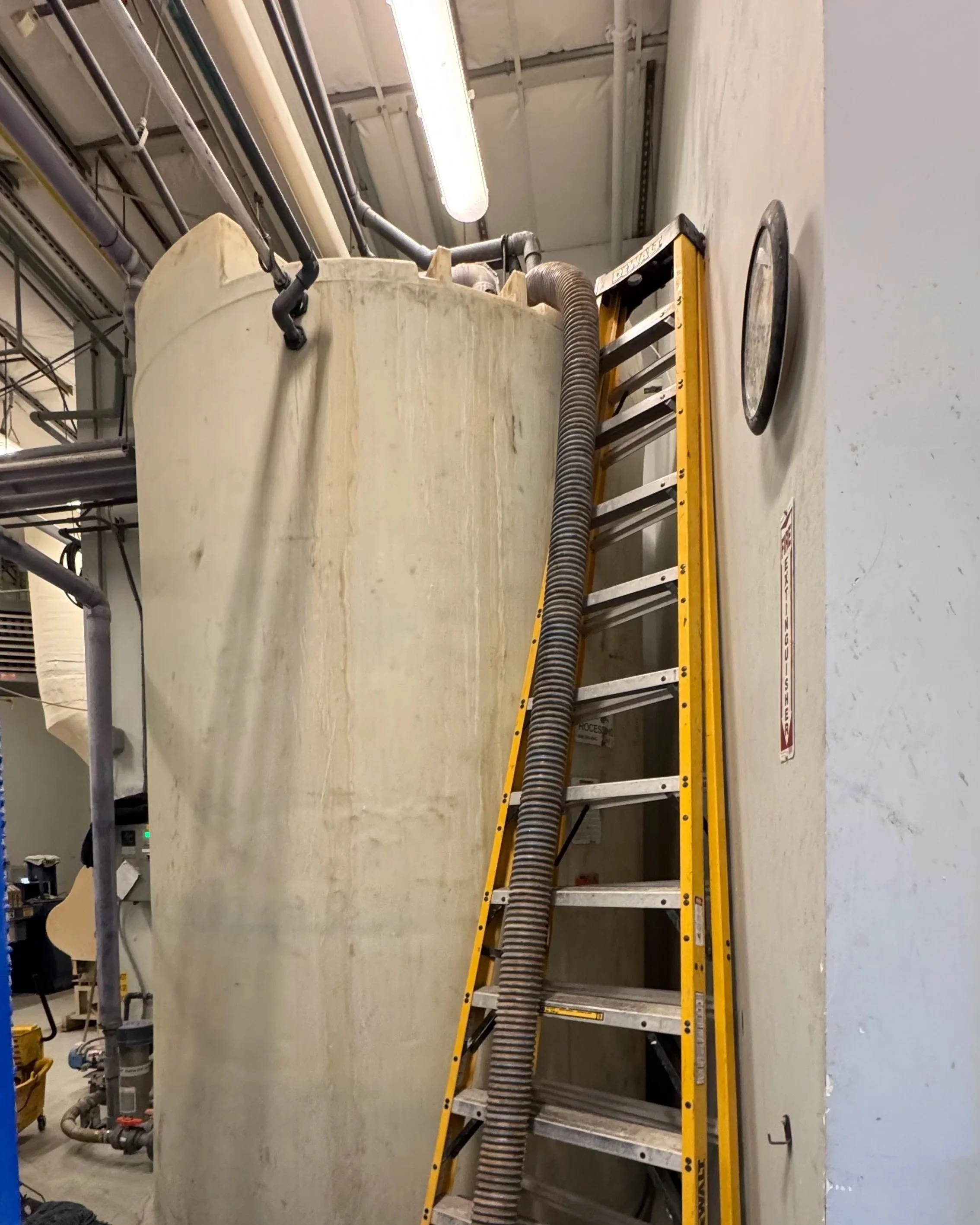 A large industrial tank with a yellow ladder leaning against it, a flexible hose connected at the top, and concrete spill or wet area at the base of the tank.