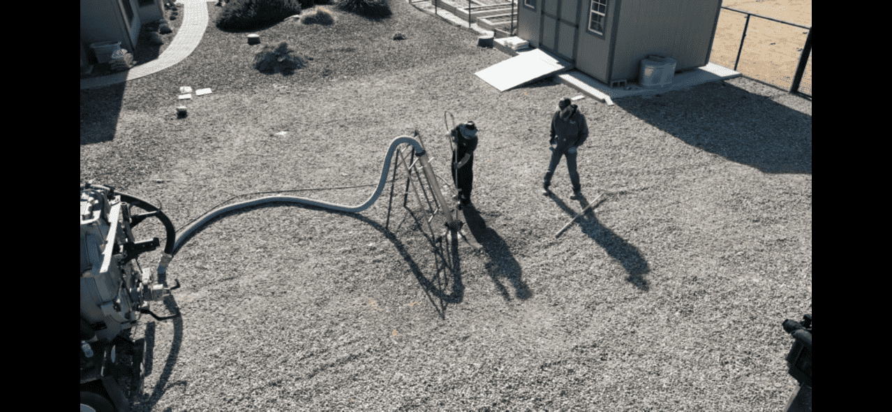 Two people walking in a gravel playground area near a small building with a ramp, surrounded by a fence, with playground equipment nearby and shadows cast on the ground.
