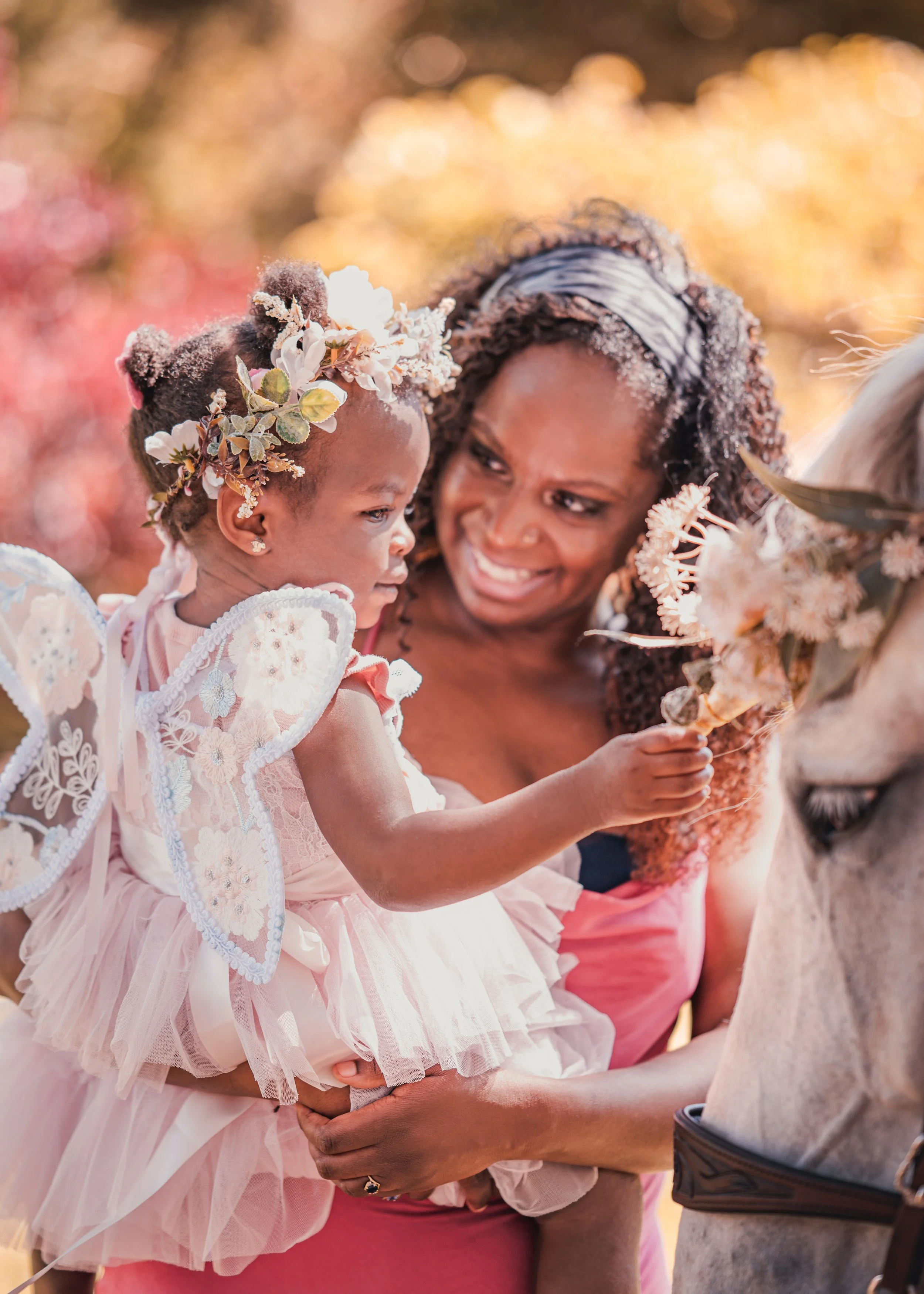 A little girl dressed in a pink fairy costume with wings is held by a smiling woman. The girl is offering flowers to a horse. The background is a sunny, blurred garden.