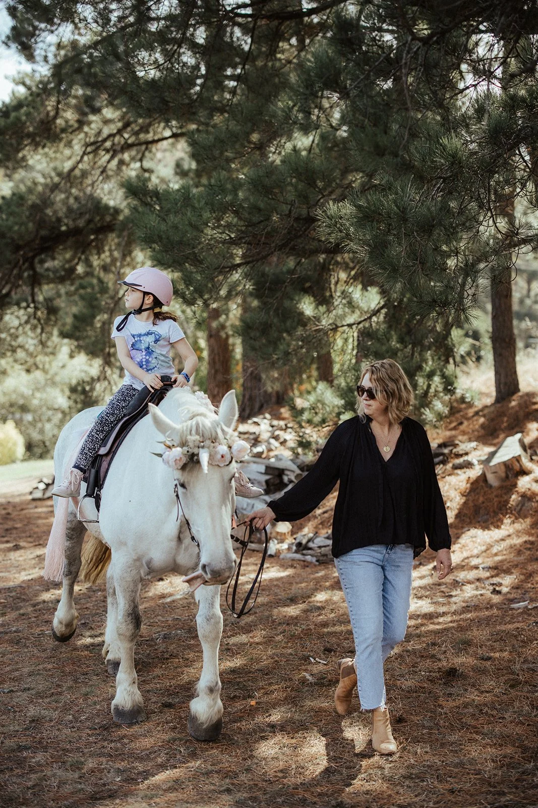 Child riding a white horse with unicorn decorations, led by a woman in a wooded area.