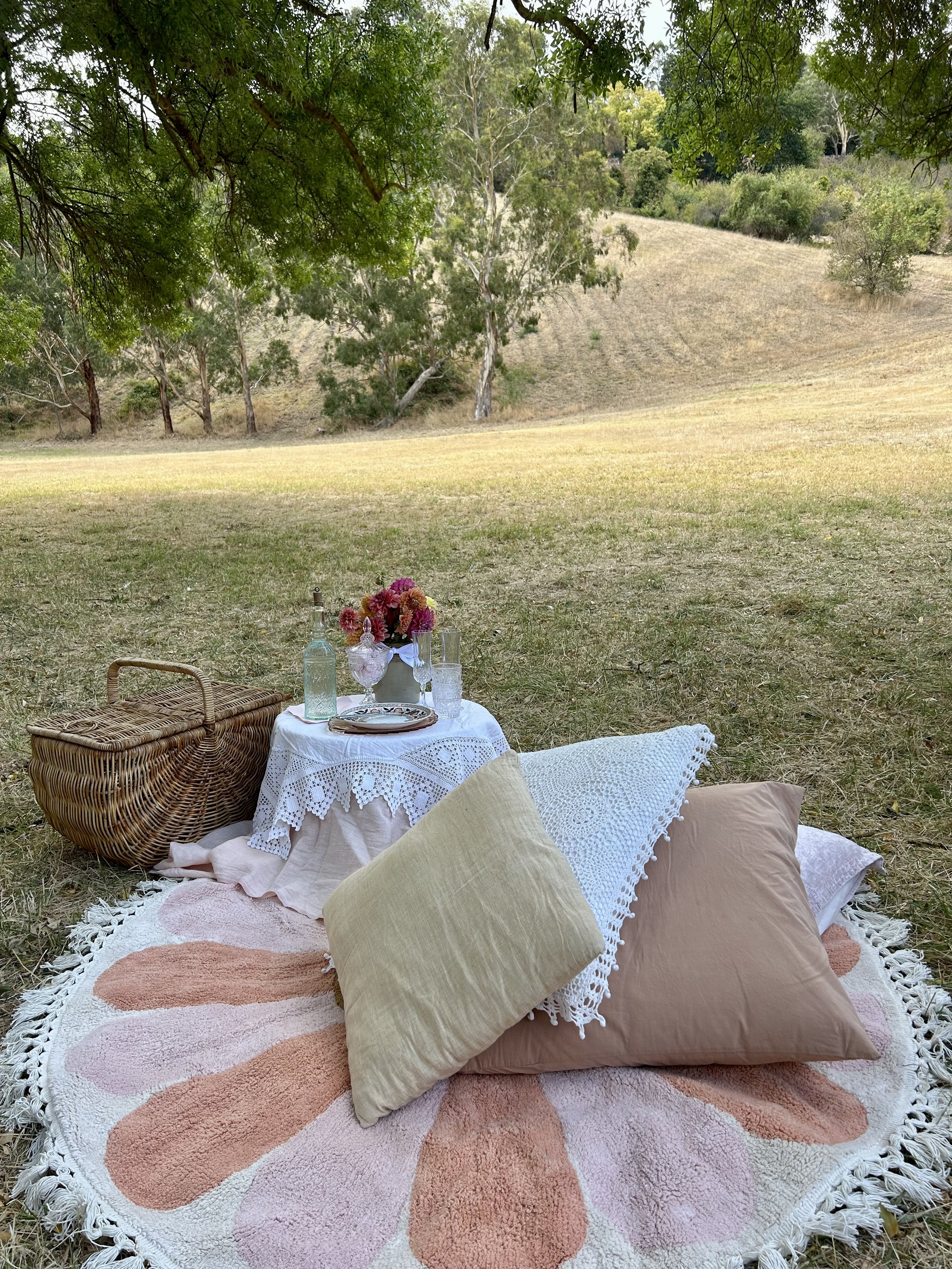 Outdoor picnic setup with a floral-patterned round rug, cushions, a small table with lace cloth holding flowers and drinks, a wicker basket, and a grassy field under trees.