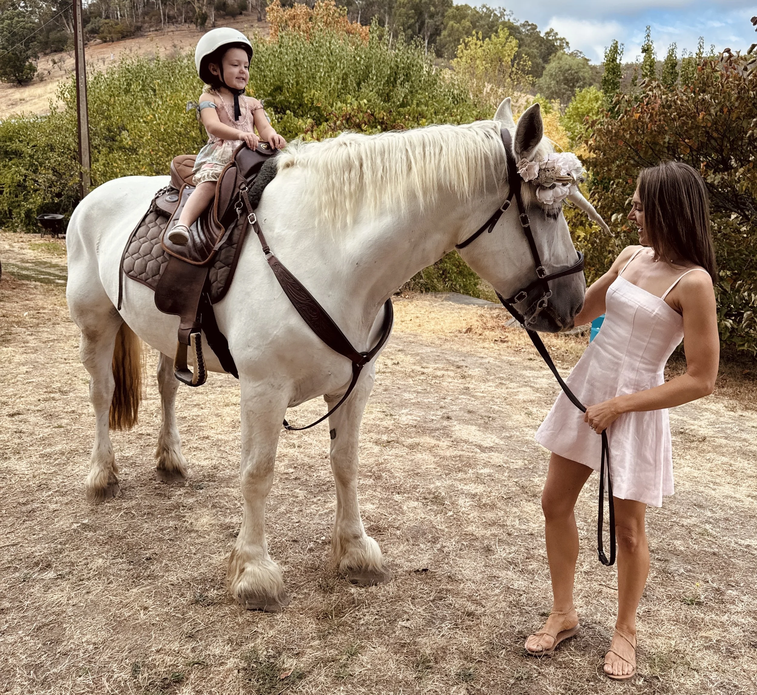 A young child wearing a helmet is sitting on a white horse that is decorated with a unicorn horn and a floral headband. An adult in a light sundress is holding the horse's reins, standing beside it. The setting appears to be an outdoor area with tree