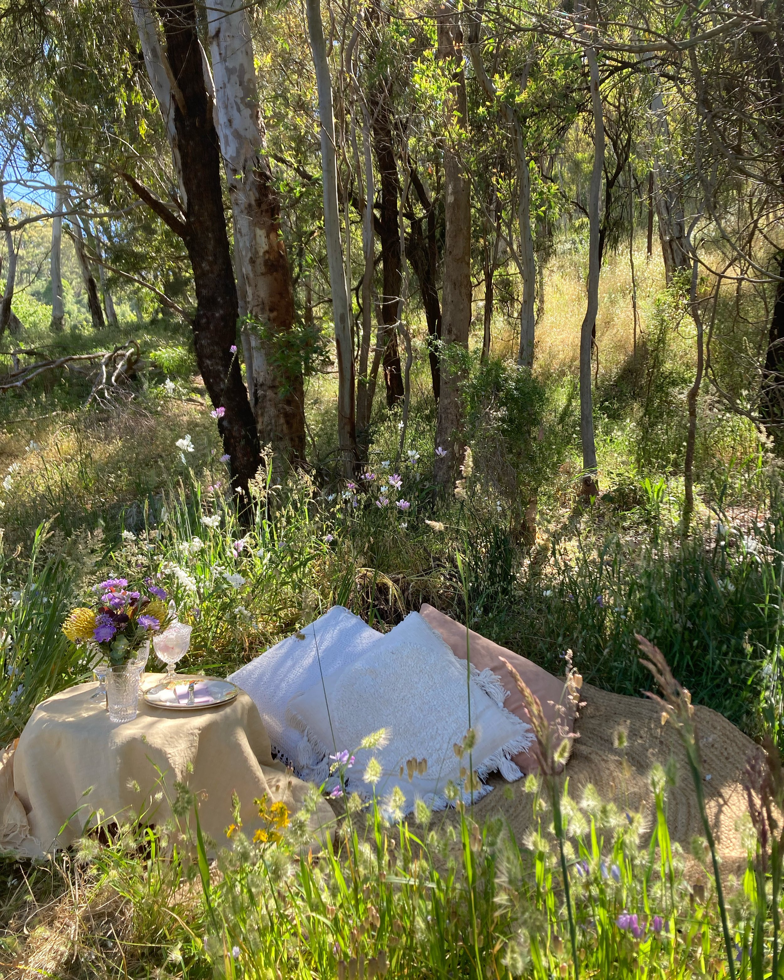 A rustic picnic setup in a forest clearing with cushions, a table with a beige cloth, a vase of wildflowers, and two glasses.