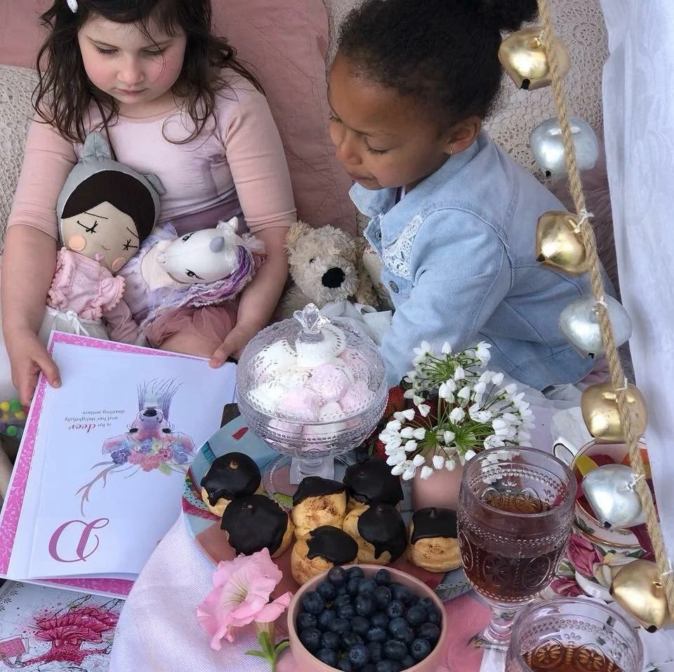 Two children having a tea party with dolls and stuffed animals, surrounded by desserts including chocolate-covered pastries, blueberries, and pink sweets, with flowers and decorative lights.