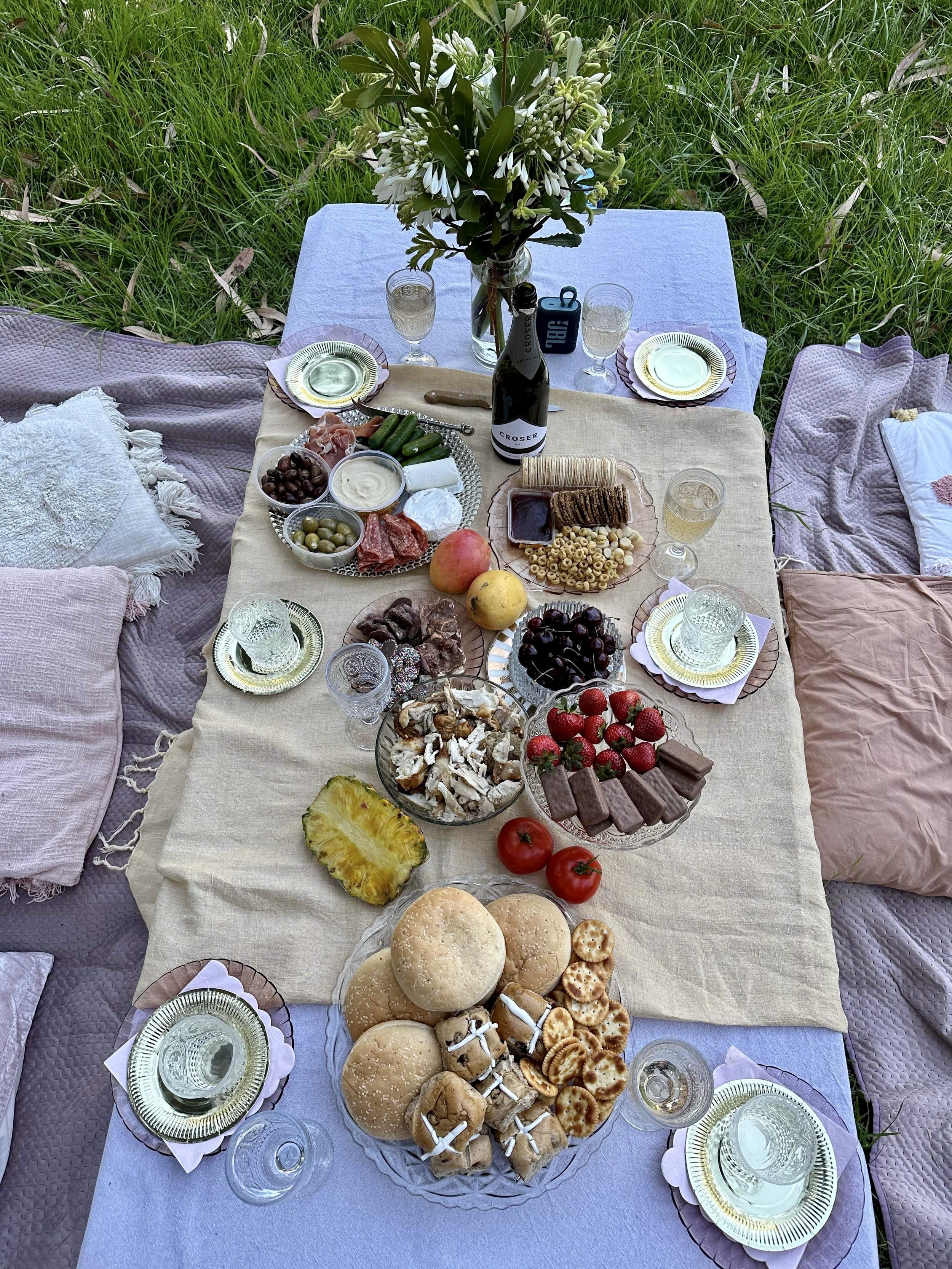 Outdoor picnic setup with a tablecloth on grass, featuring plates of fruits, vegetables, cheeses, crackers, bread rolls, and pastries. A wine bottle, drinking glasses, and a vase of flowers are also present.