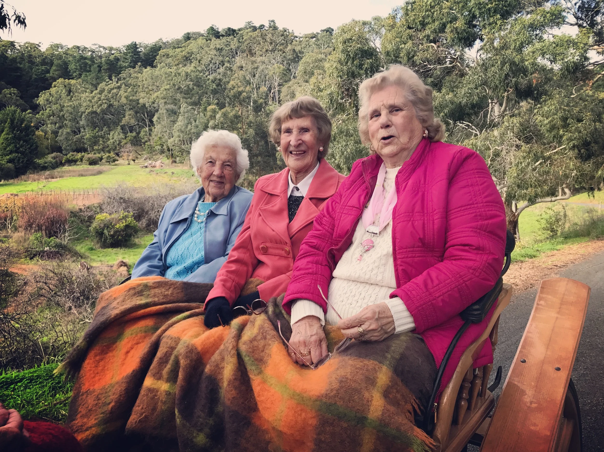 Three elderly women sitting on a bench outdoors, smiling and covered with a blanket, surrounded by trees and greenery.