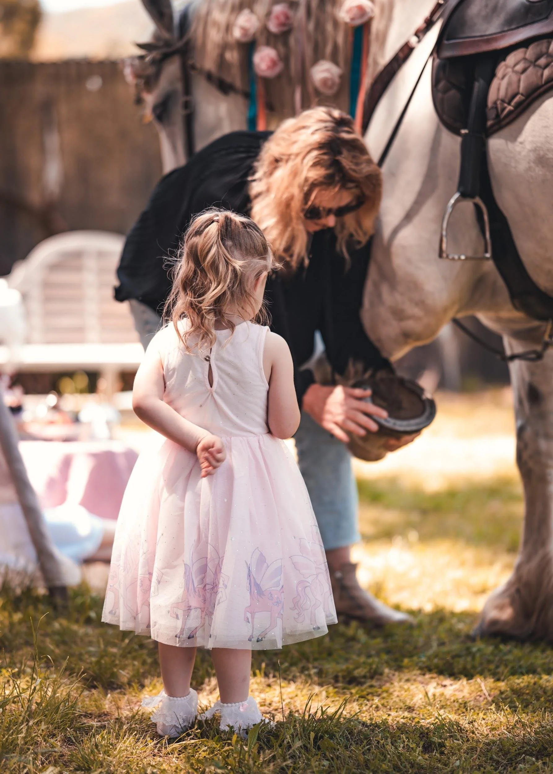 Young girl in a pink dress watching a woman tending to a horse's hoof outdoors