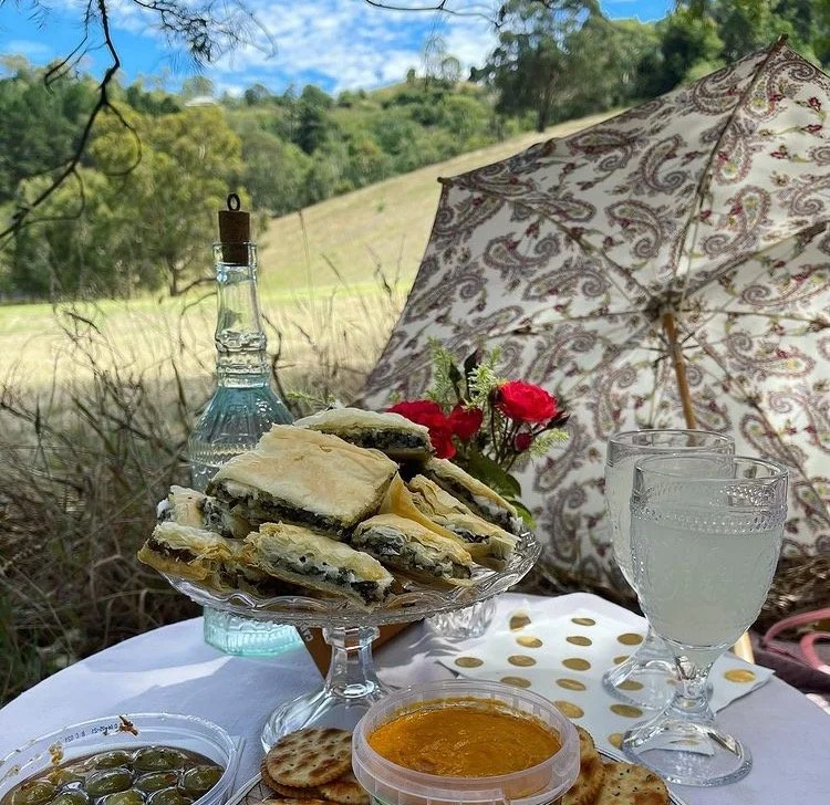 Picnic setup with pastries and drinks on a table, decorative umbrella, greens and flowers, outdoor scenery