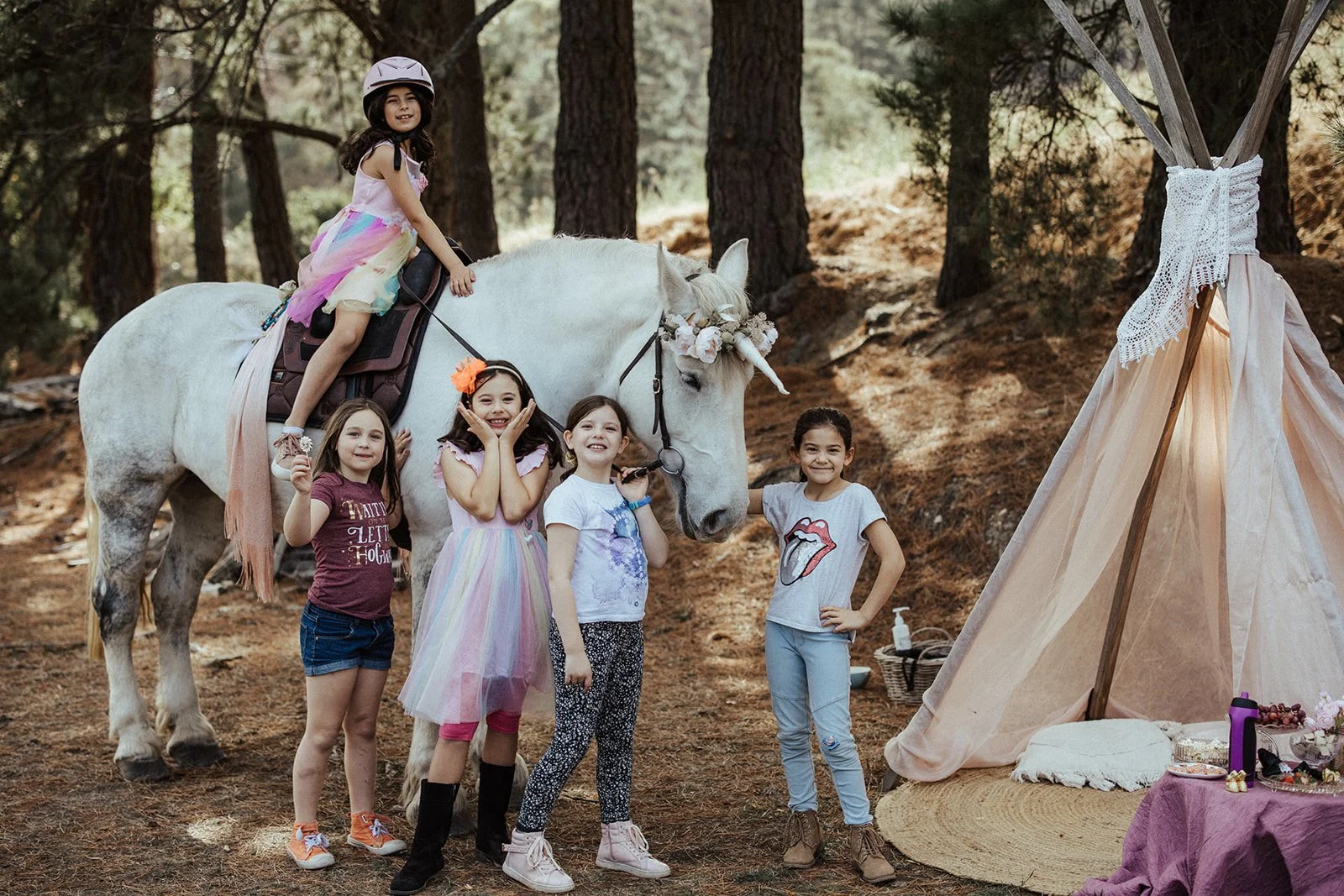 Group of children posing with a white horse dressed as a unicorn next to a tent in a forest setting.