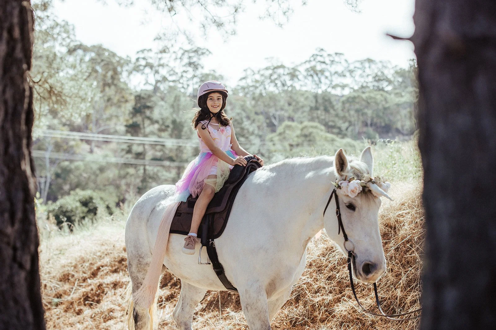 A child wearing a helmet and a colorful tutu rides a white horse with a unicorn horn and floral decorations in an outdoor setting.