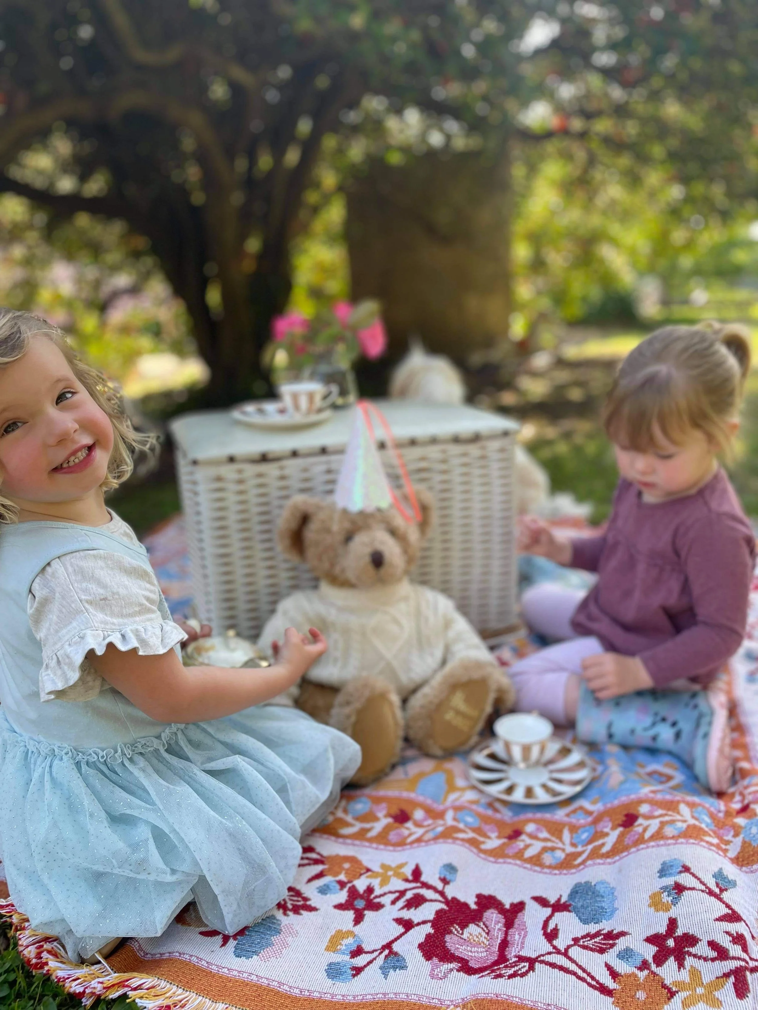 Two young children having a tea party outdoors on a colorful blanket with a teddy bear wearing a sweater. The children are sitting near a wicker picnic basket. The scene is under a tree, surrounded by greenery.