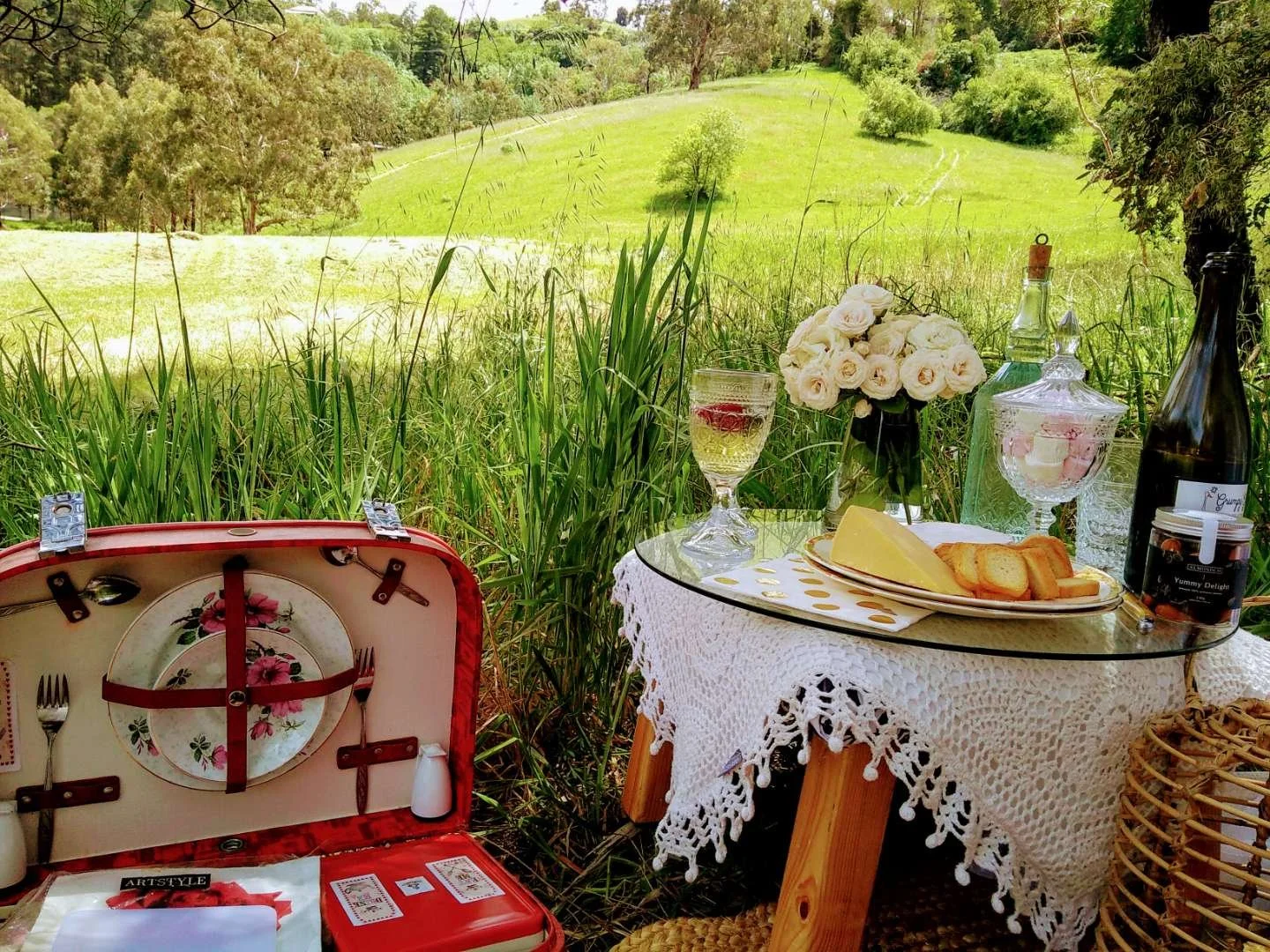 Outdoor picnic setup with a packed picnic basket, cheese plate, wine glasses, and white roses on a small table, surrounded by lush greenery.