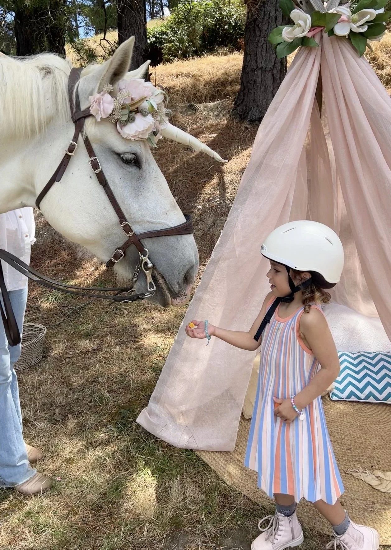 A child wearing a helmet feeds a white horse with a unicorn horn and floral headpiece near a cloth tent in a wooded area.
