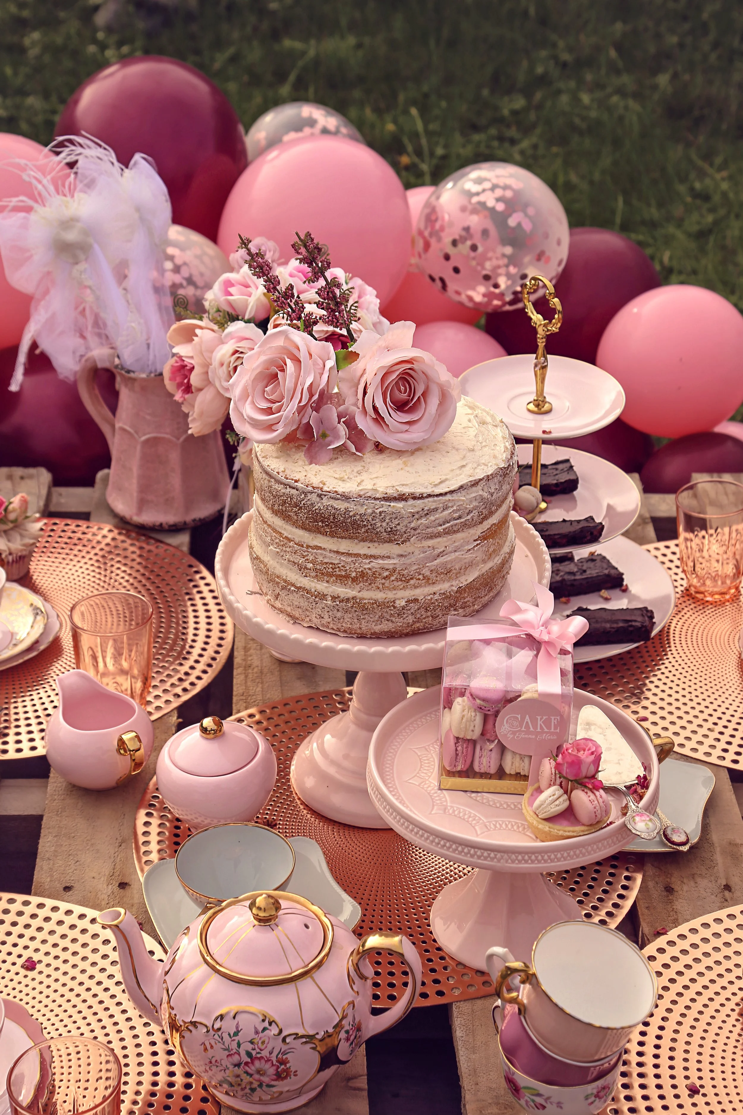 Outdoor tea party table setting with a naked cake adorned with pink flowers, surrounded by pink and burgundy balloons, pastel teapots, cups, macarons, and decorative plates.