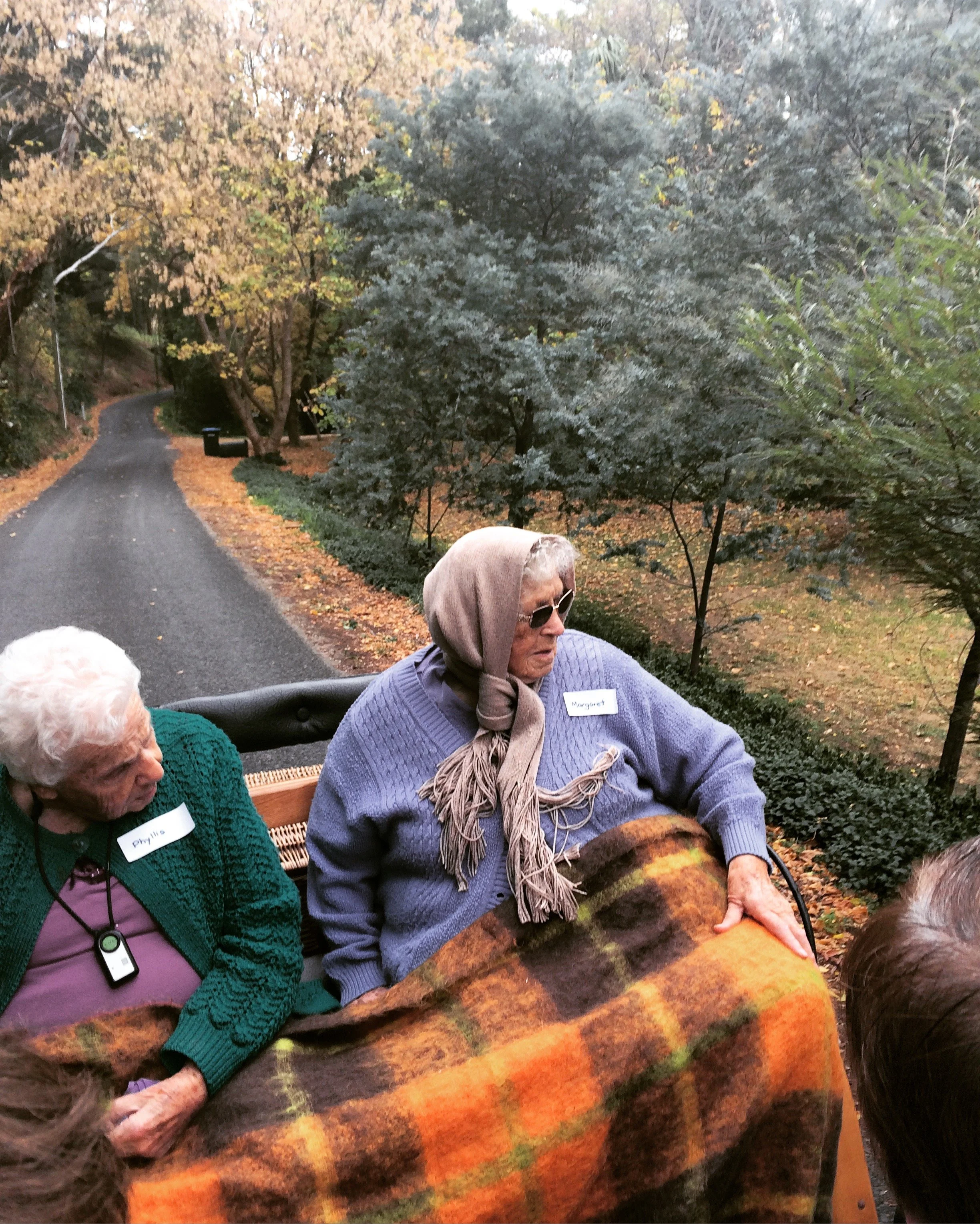 Elderly people in a wooded area, sitting on a vehicle with a plaid blanket, wearing coats and headscarves.