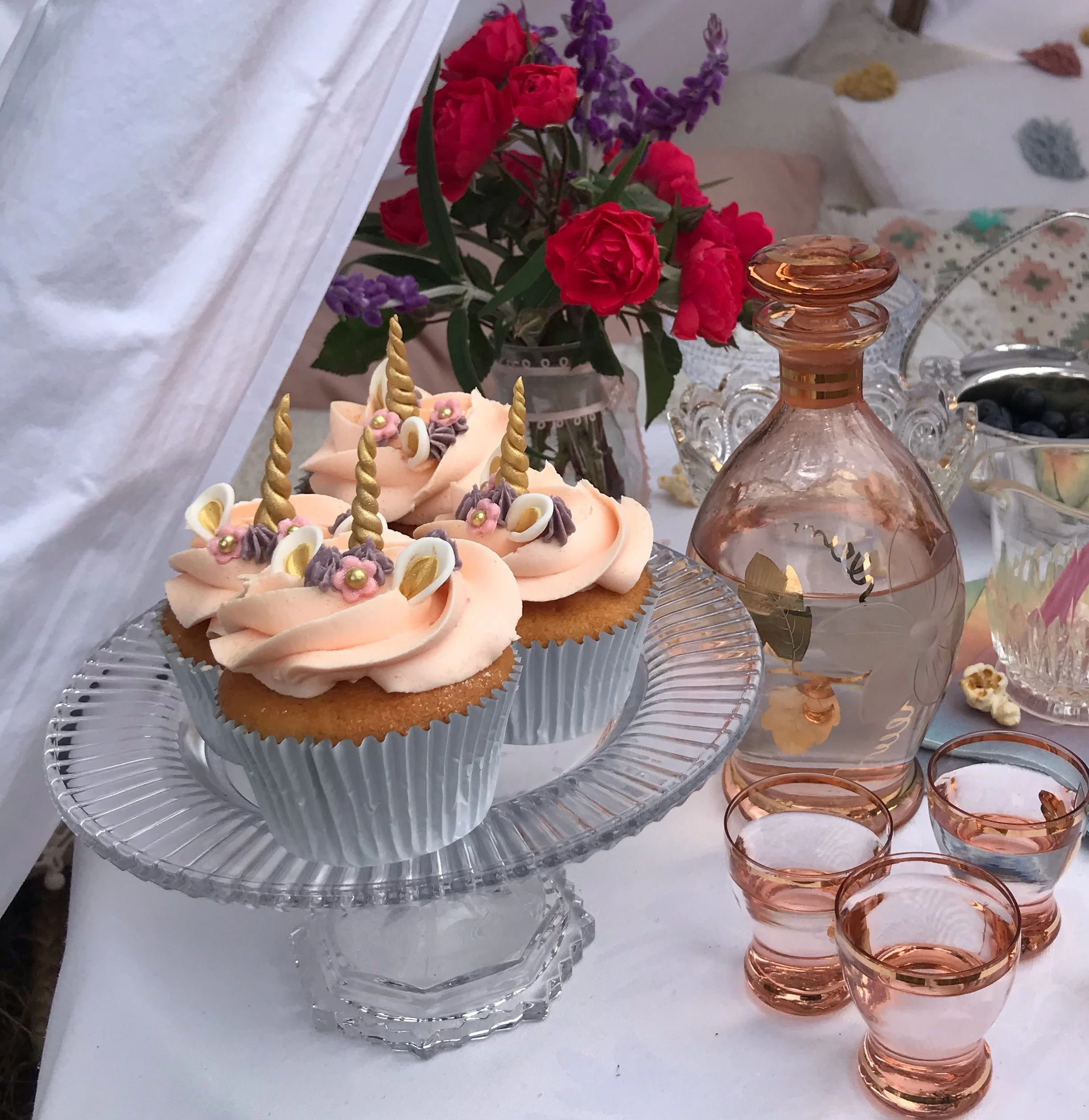 Cupcakes with unicorn decorations on a glass stand, a crystal decanter, and glasses on a table beside red and purple flowers.