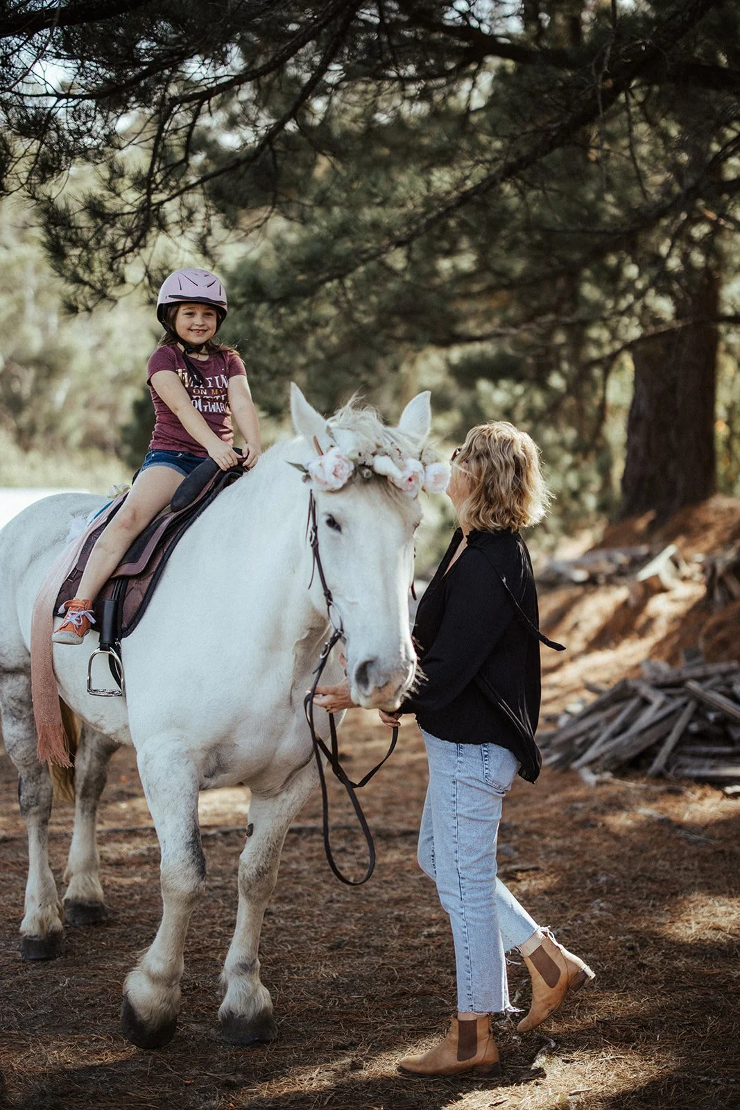 Child sitting on a white horse with a floral wreath, guided by an adult in a forest setting.