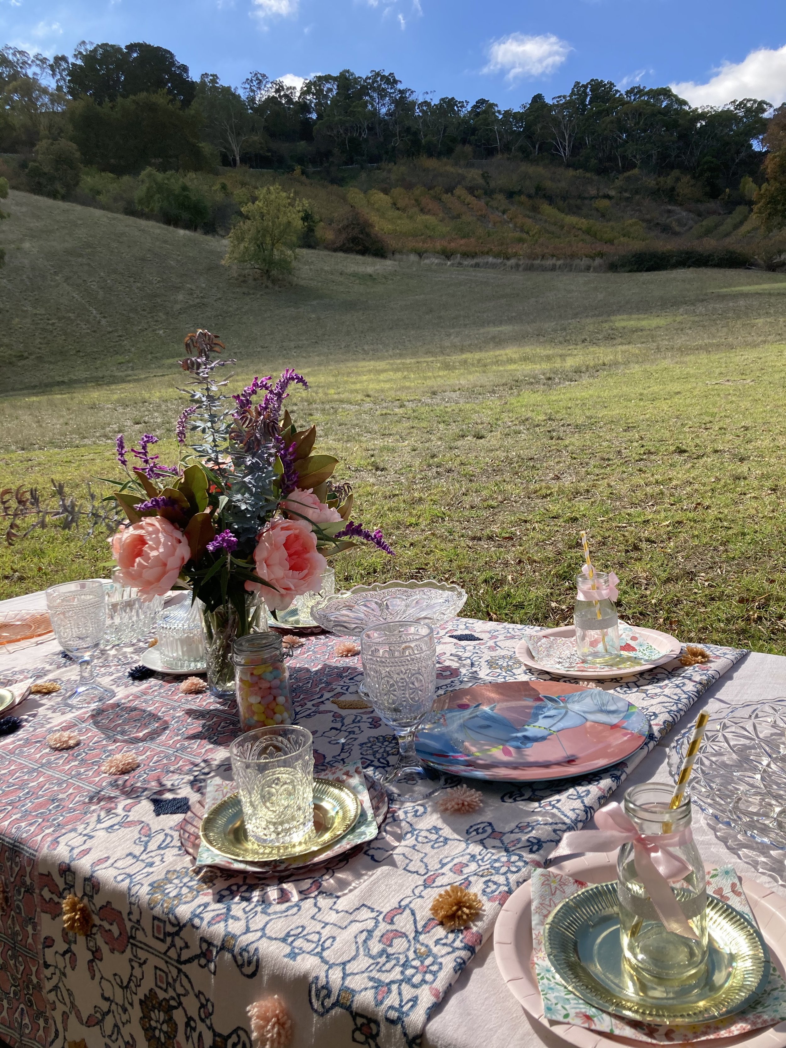 A table set for an outdoor picnic with floral decorations and glassware, overlooking a grassy field and wooded hills.