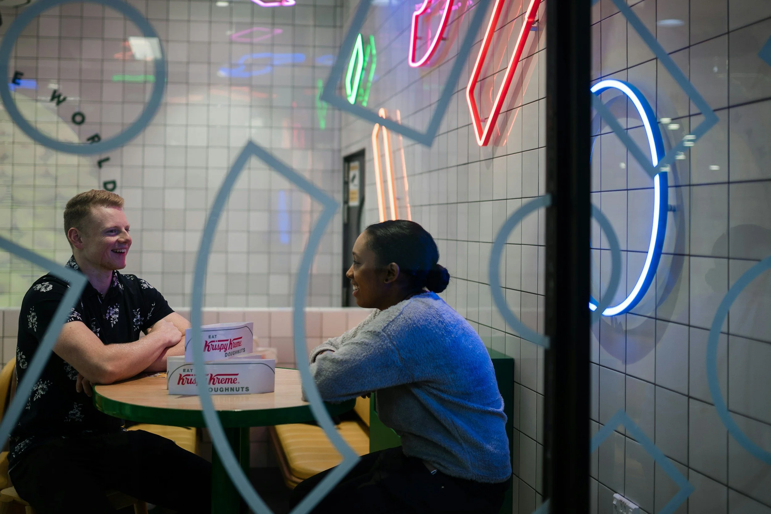 Two people sitting at a table with Krispy Kreme doughnut boxes in a cafe with colorful neon signs and tiled walls.