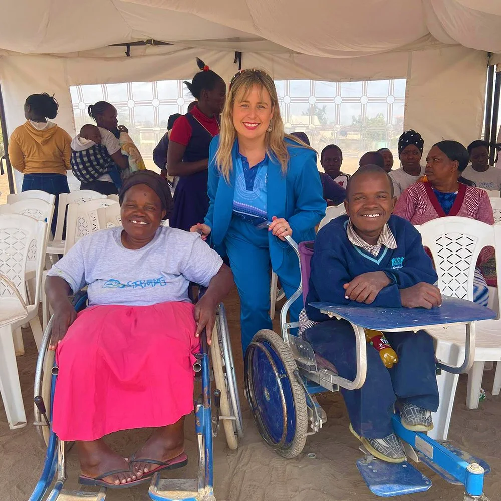 Mama Respond Founder Erin Hagerty - Photographed as a group in a tent, with two women in wheelchairs that the charity supports, Erin is the smiling woman standing between them with others seated and standing in the background.