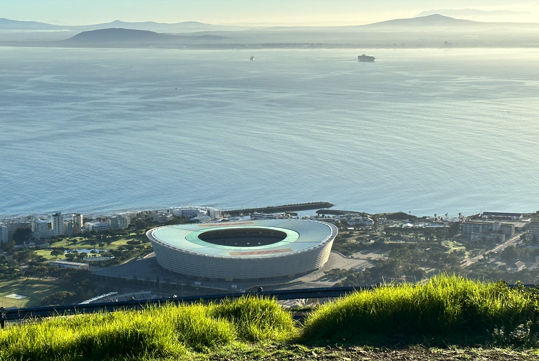 Aerial view of a stadium near the waterfront with surrounding buildings, green spaces, and boats on the water, with hills in the background.
