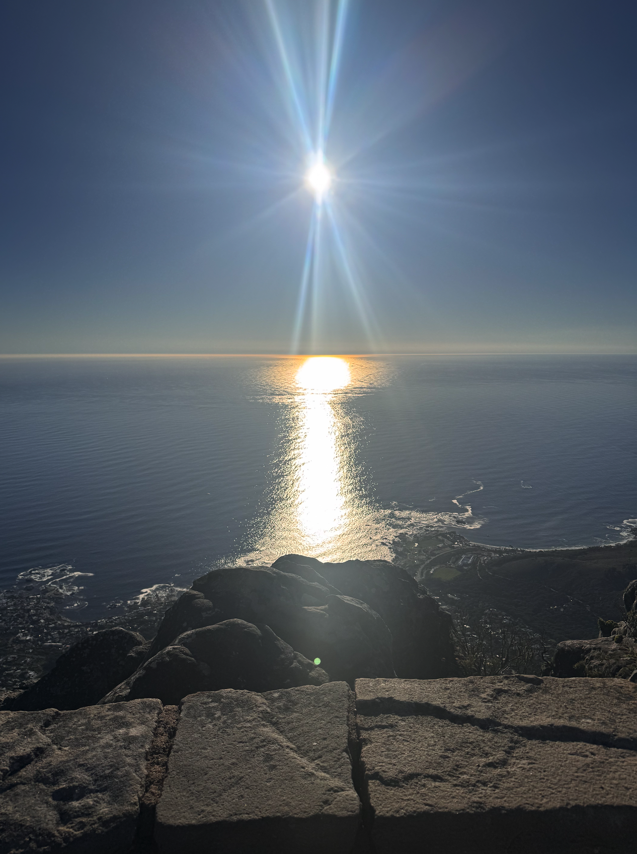 View from Table Mountain over the ocean with the sun shining brightly, creating a reflection on the water, and rocks in the foreground.