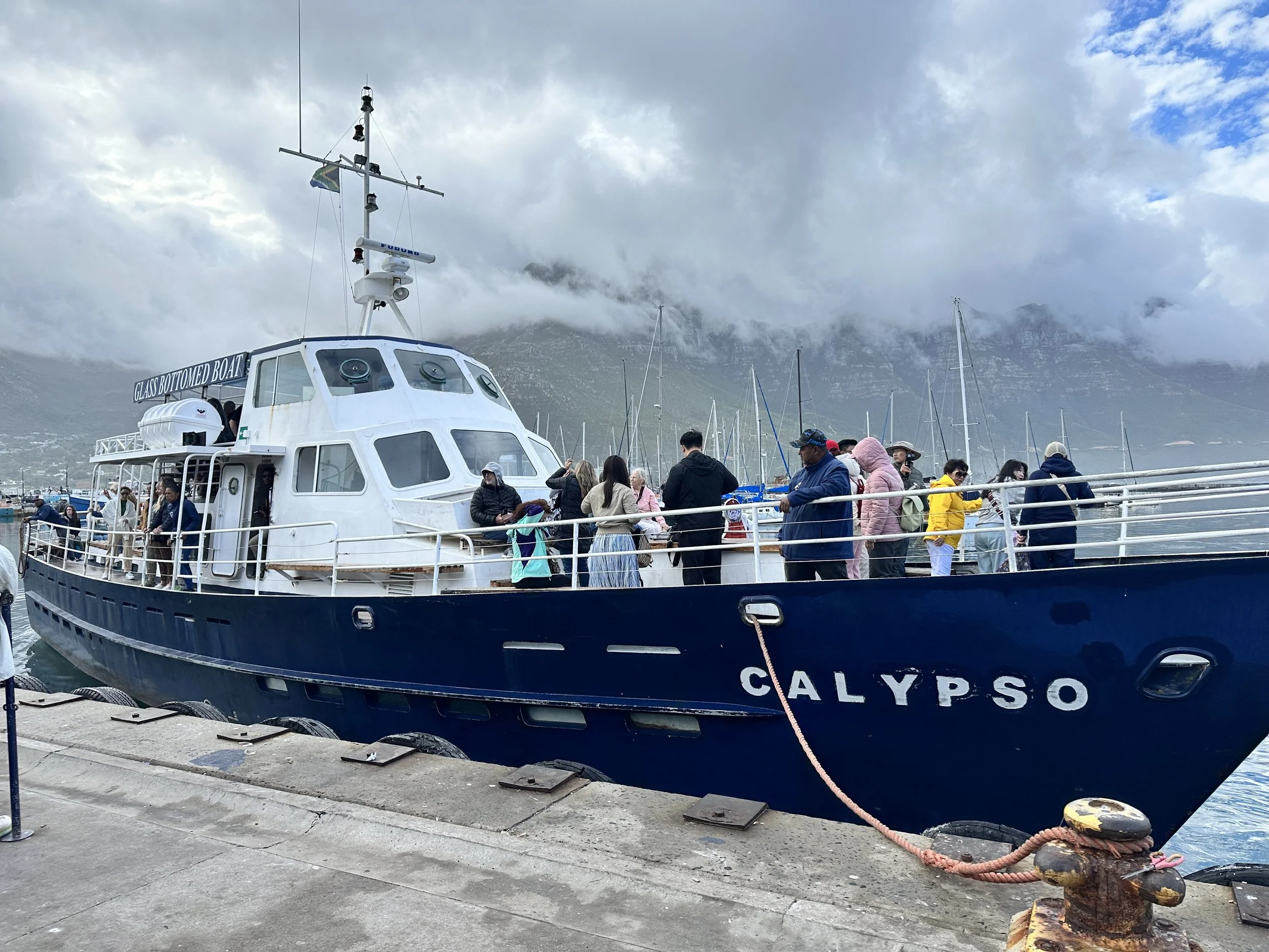 A boat named CALYPSO docked at a marina with people on board and a mountain in the background.
