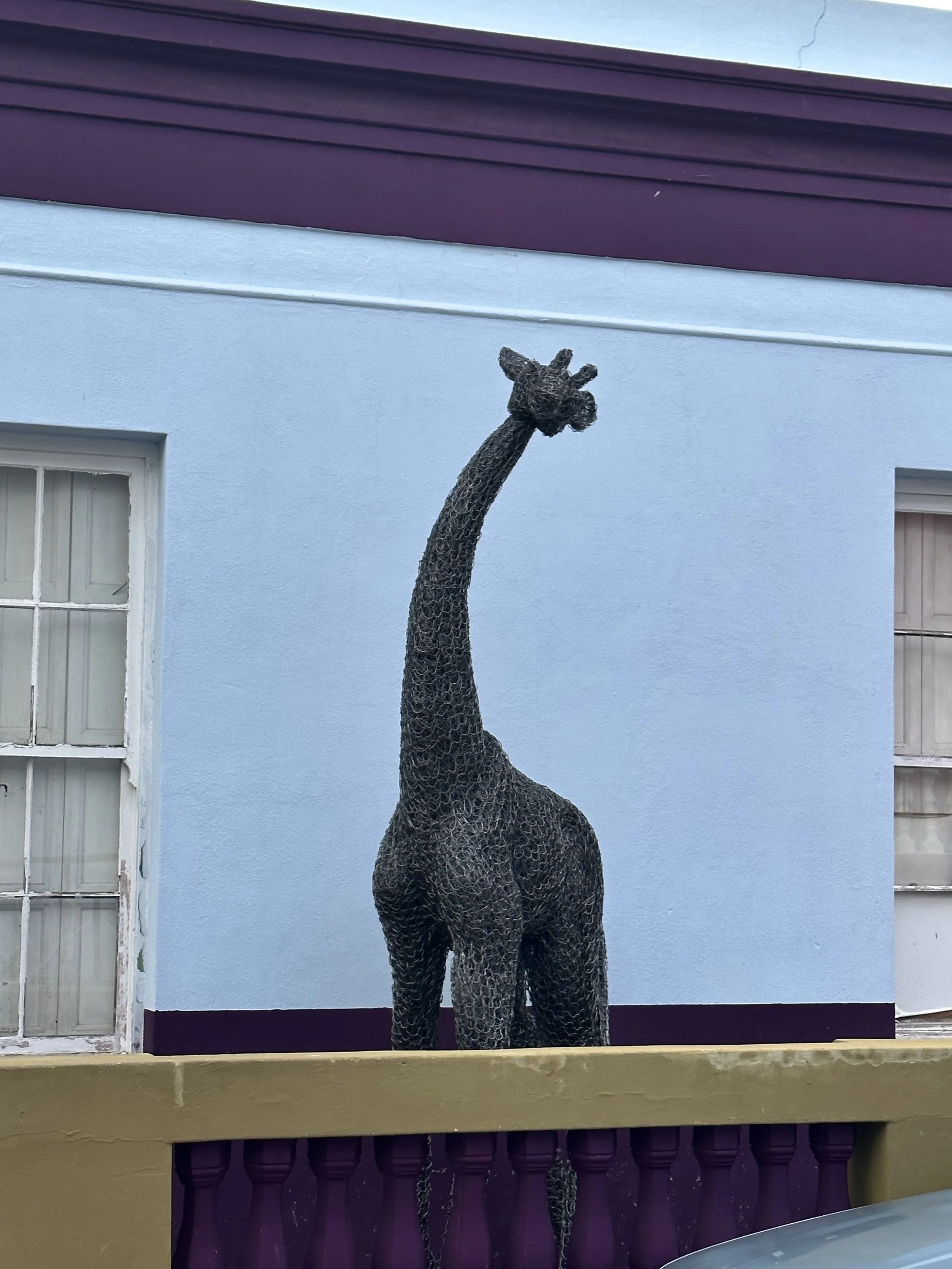A large sculpture of a giraffe made from dark metal mesh stands outdoors against a light blue wall with purple trim.
