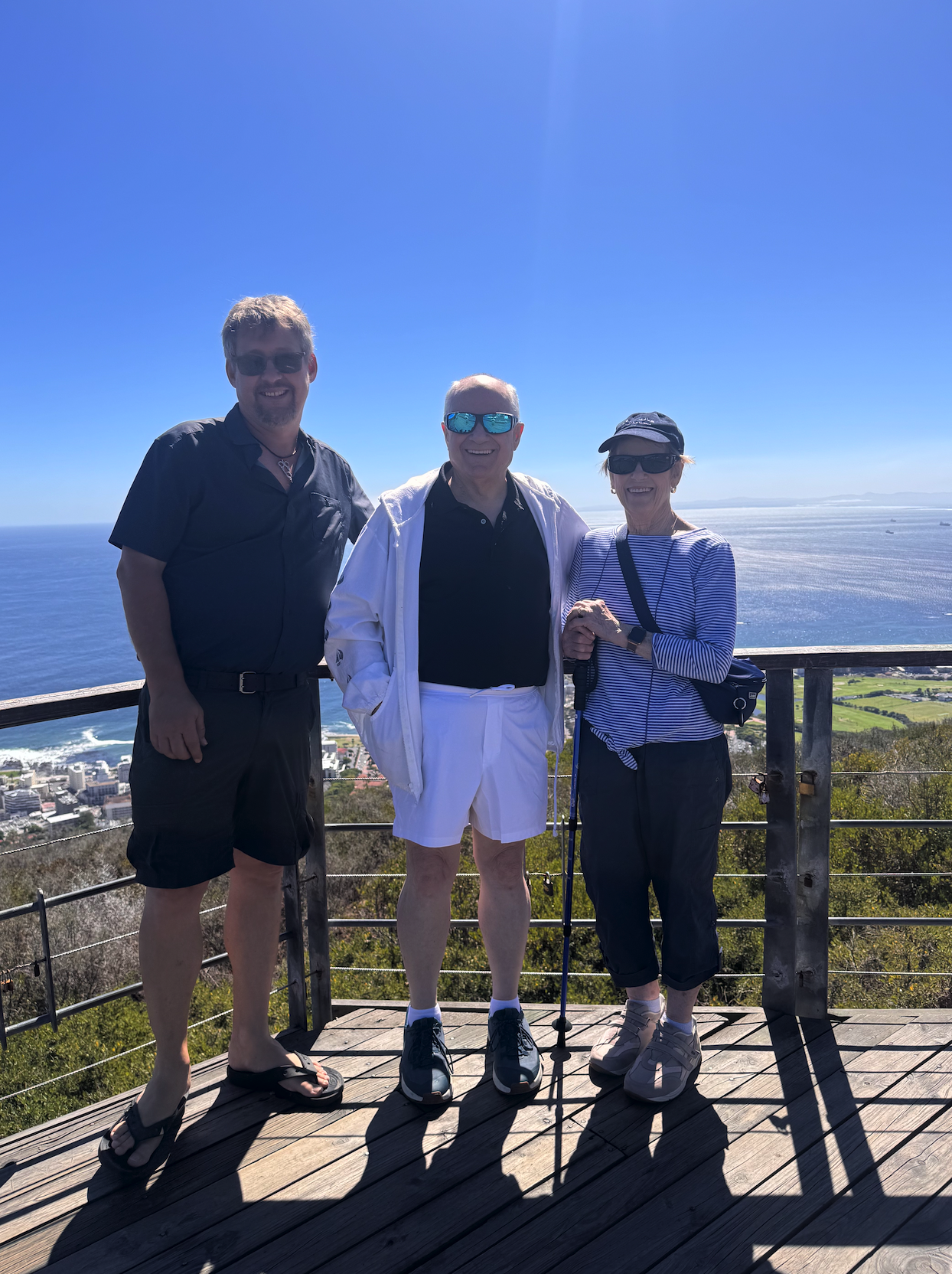 Three people standing on a wooden viewing platform with the ocean and coastline behind them, smiling at the camera on a sunny day.