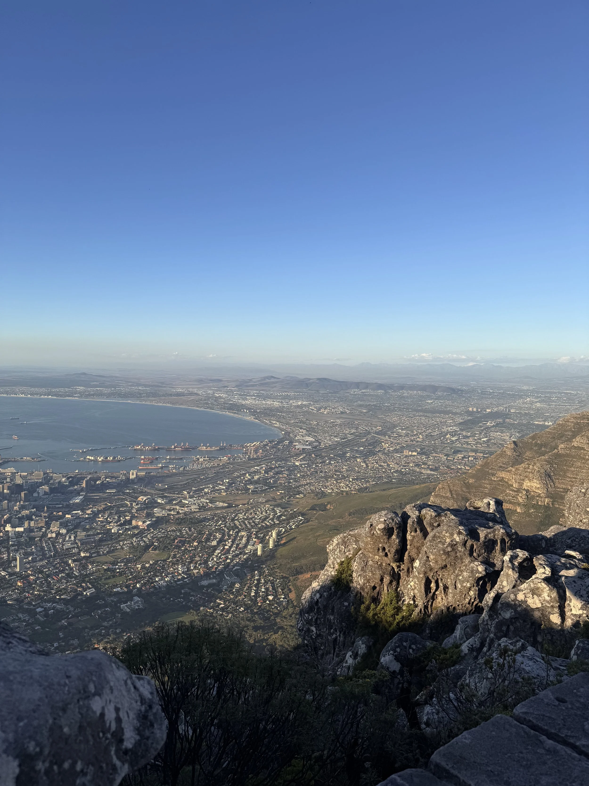 A panoramic view of a city next to a large body of water, seen from a mountain or hill, with rocks and sparse vegetation in the foreground and a clear blue sky above.