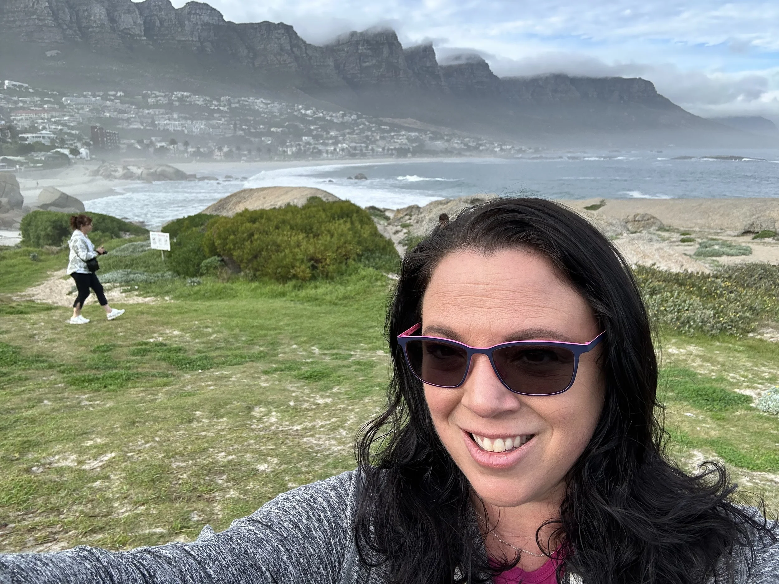 A woman with long black hair wearing sunglasses and a gray jacket taking a selfie at the beach with green grass, large rocks, and ocean waves in the background. A person in white walking along a dirt path, and cliffs or mountains with clouds overhead