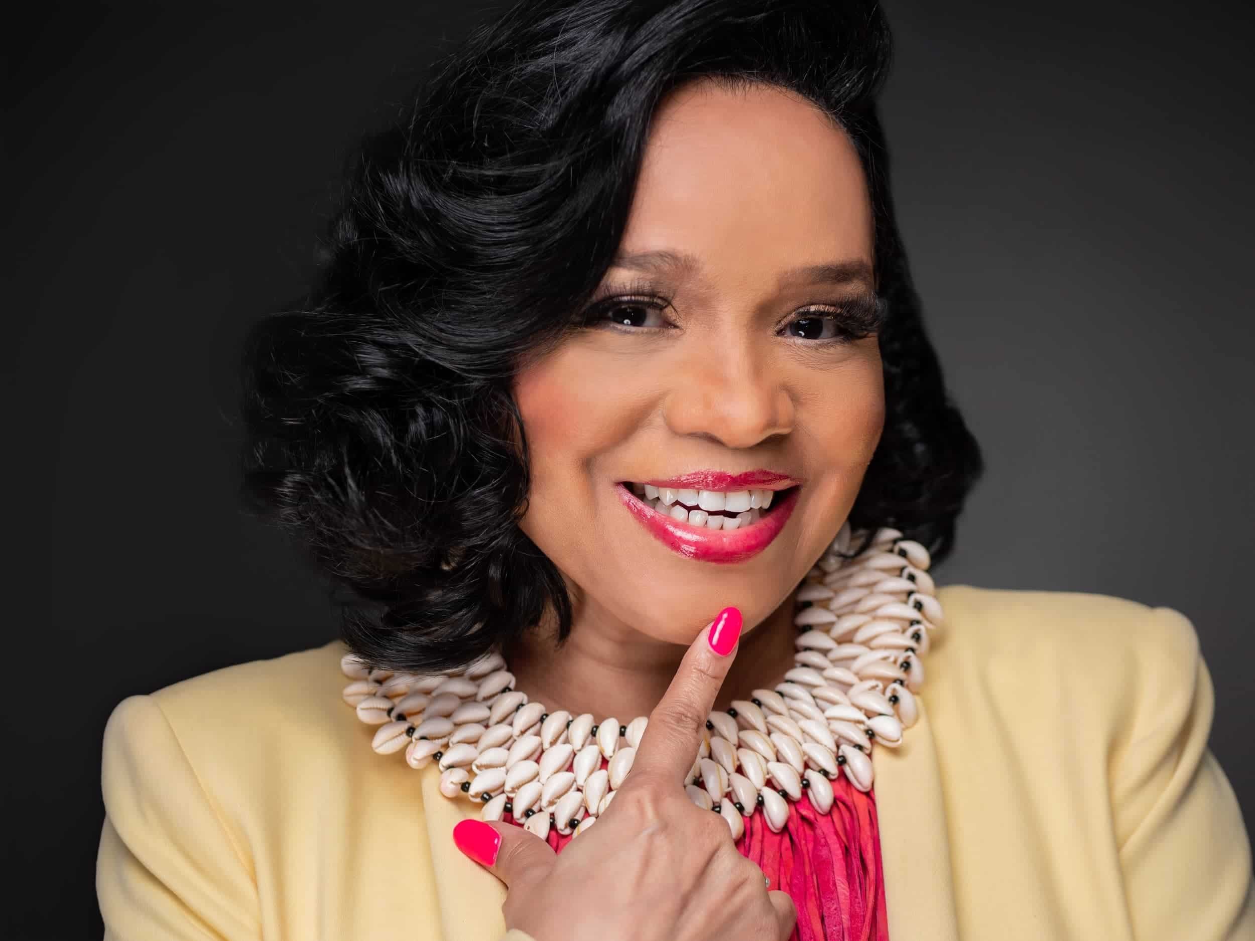 A smiling woman with dark, curly hair in a professional portrait. She is wearing a yellow blazer, a white shell necklace, a pink top, and bright pink lipstick, with matching pink nail polish. She is touching her chin with her right index finger against a dark background.