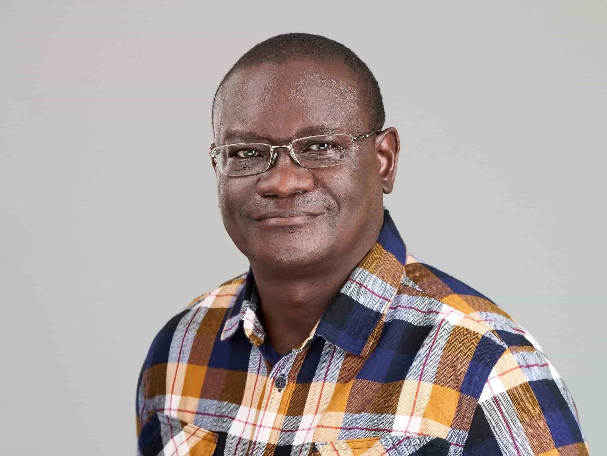 A middle-aged man with glasses wearing a multicolored plaid shirt, smiling softly, against a plain gray background.