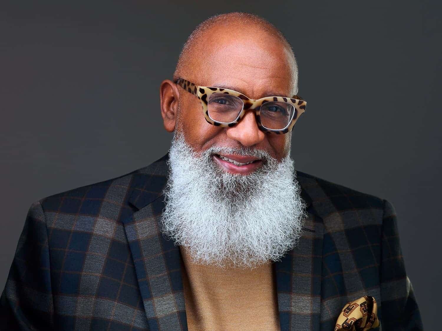 Portrait of an older man with a white beard and glasses, wearing a plaid suit and beige shirt, smiling against a dark gray background.