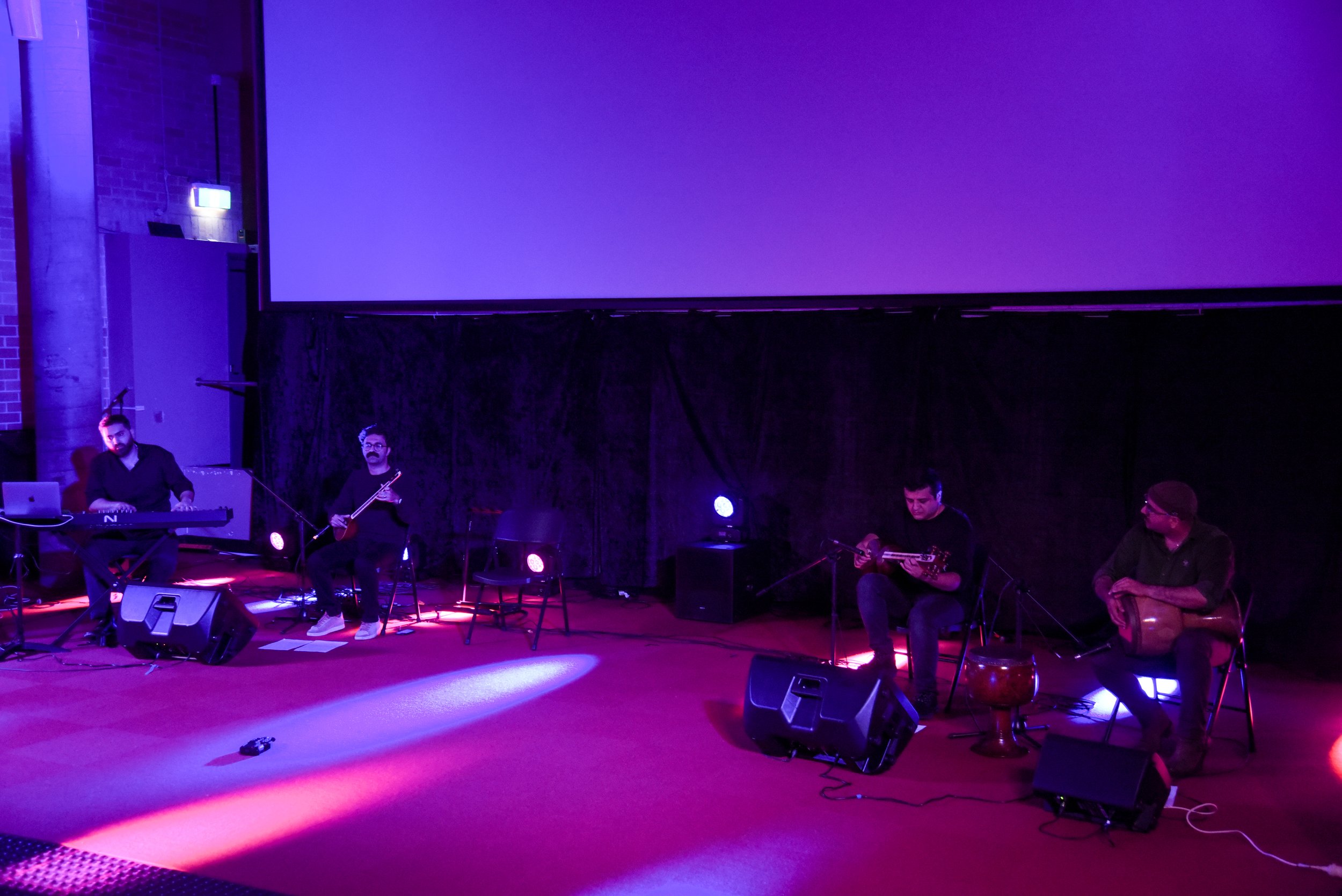 Four musicians performing on stage with purple lighting, playing keyboard, string instruments, and a percussion instrument.