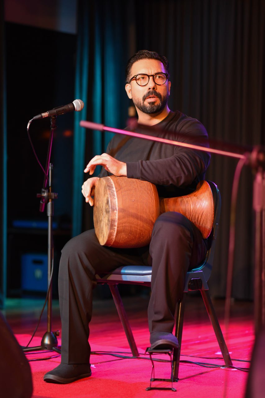 Music performance with a seated man playing a hand drum on stage with a microphone nearby.