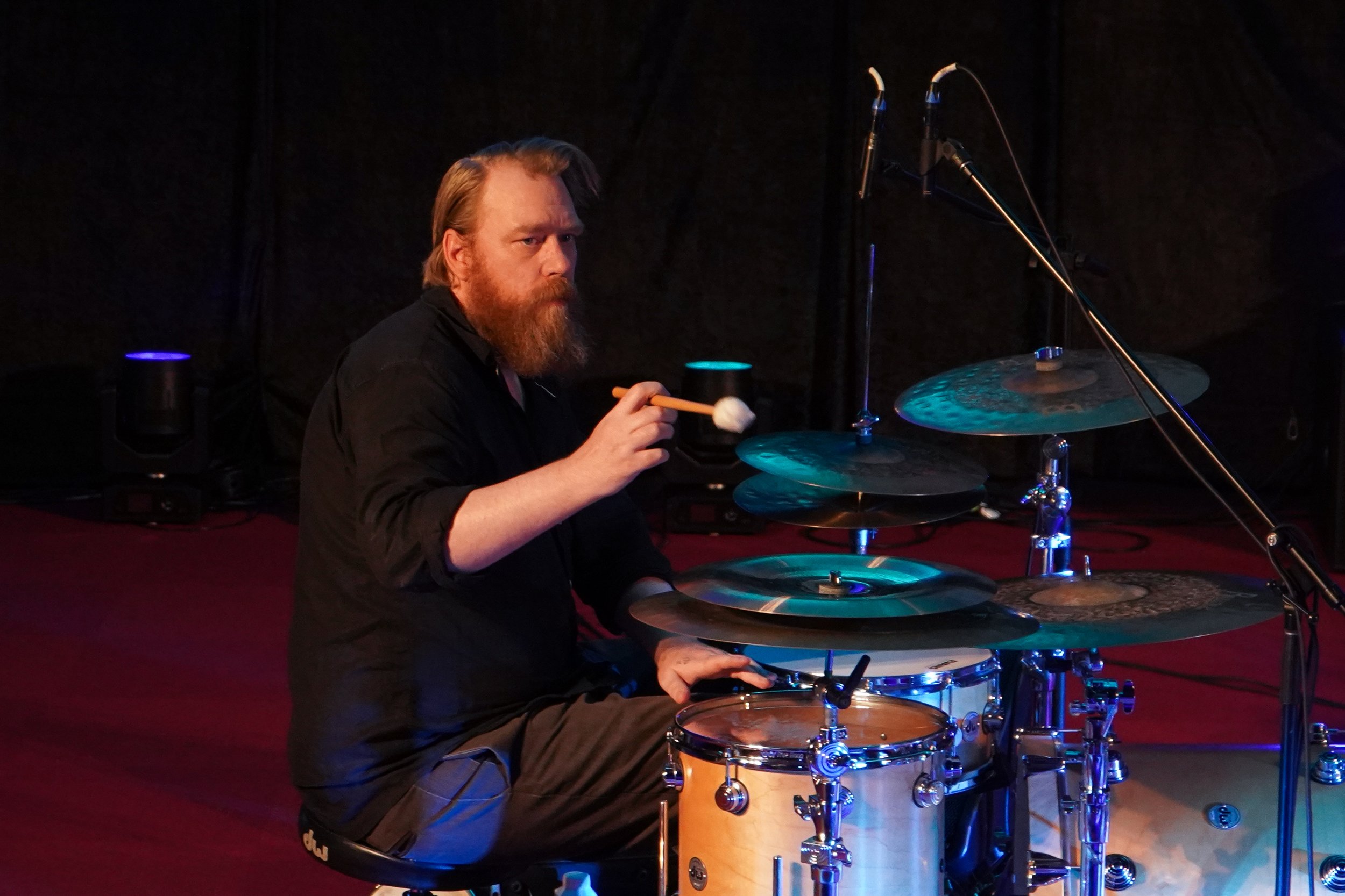 A bearded drummer performing on stage, playing a drum kit with cymbals and microphones set up.