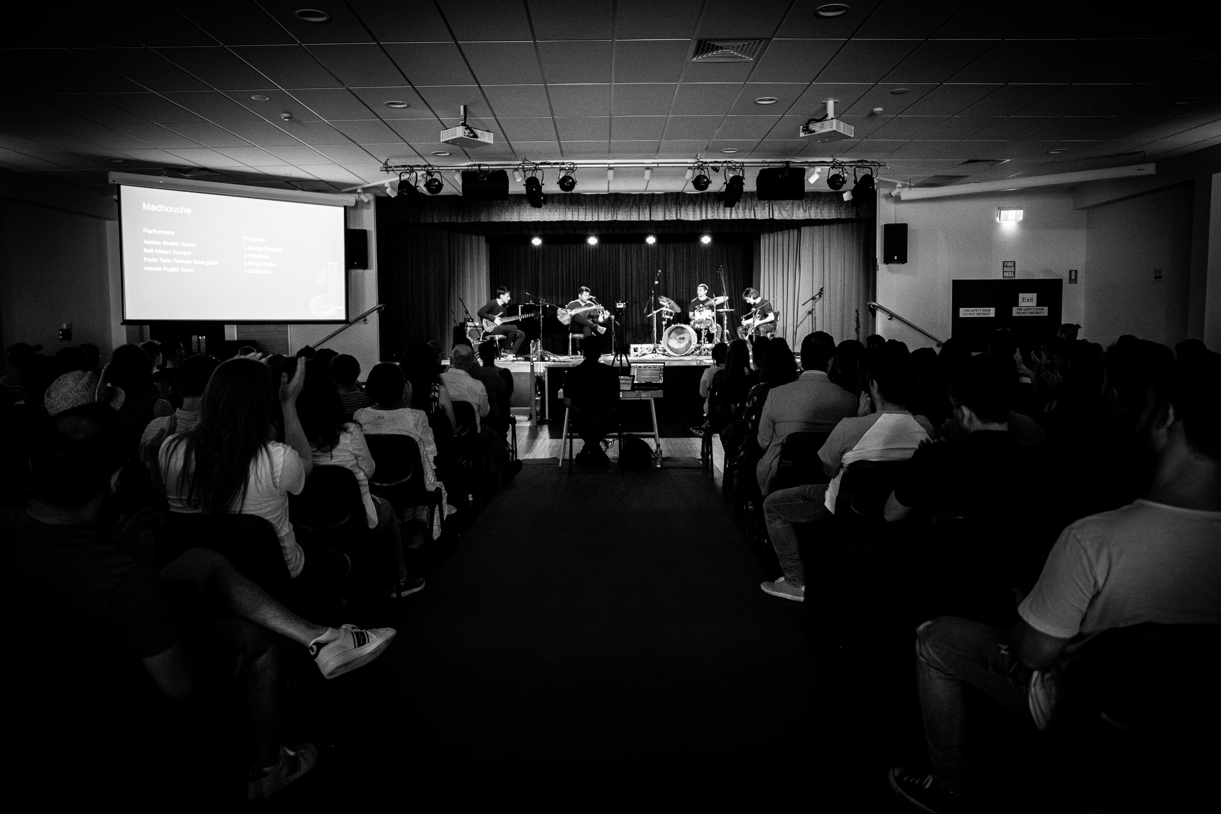 Black and white photo of a live music performance on stage in front of an audience, with a presentation screen displaying text.