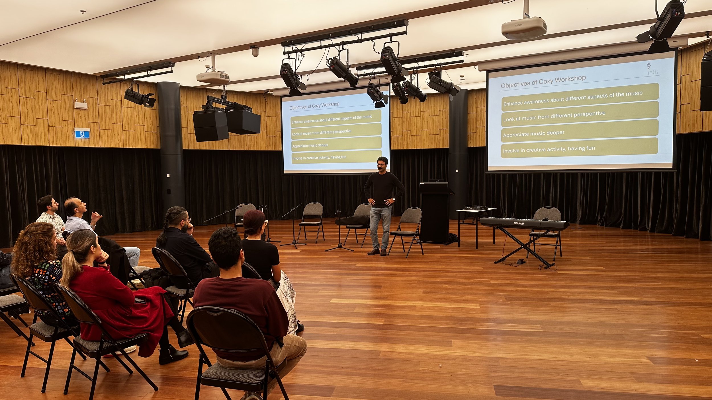 A speaker is presenting at a music workshop with an audience seated in chairs. Two projection screens display the 'Objectives of Cozy Workshop,' which include enhancing awareness about music, looking at music from different perspectives, appreciating