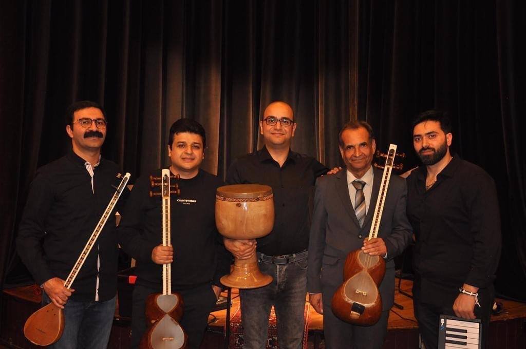 Five men standing on stage, holding traditional musical instruments, including a drum and string instruments, with a black curtain background.