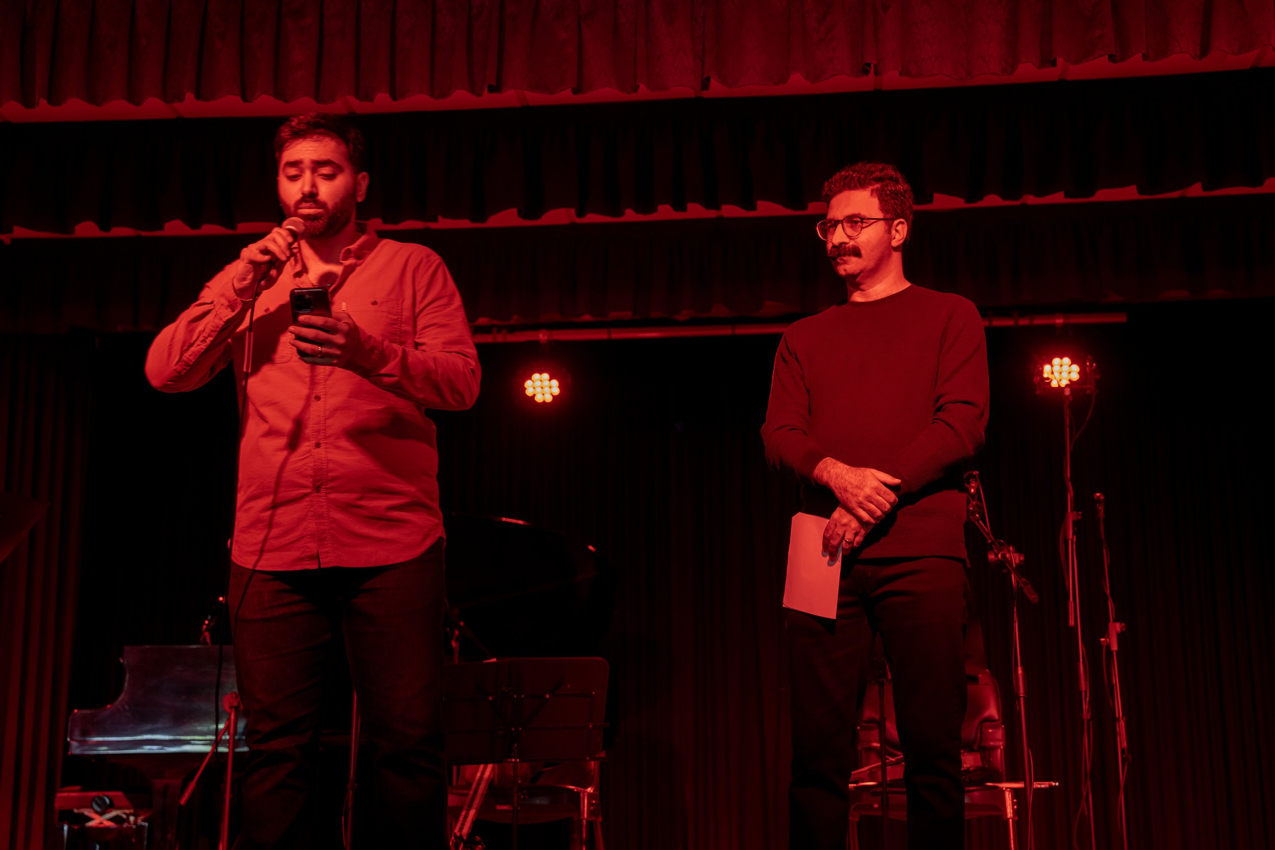 Two men on stage under red lighting, one speaking into a microphone while holding a phone, the other holding a piece of paper, with musical instruments in the background.