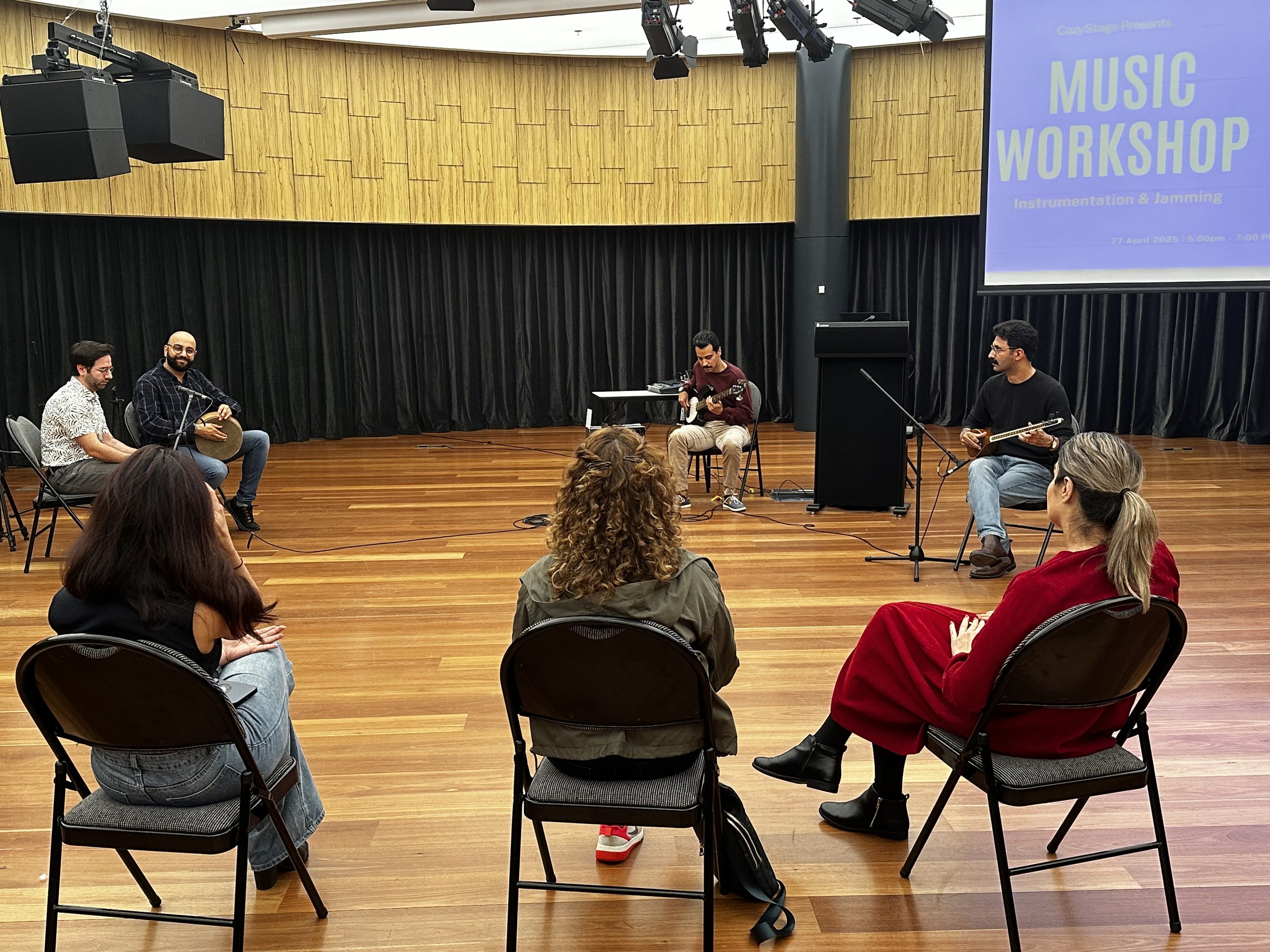 Group of people at a music workshop with musicians playing instruments on a stage, audience seated in a semi-circle.