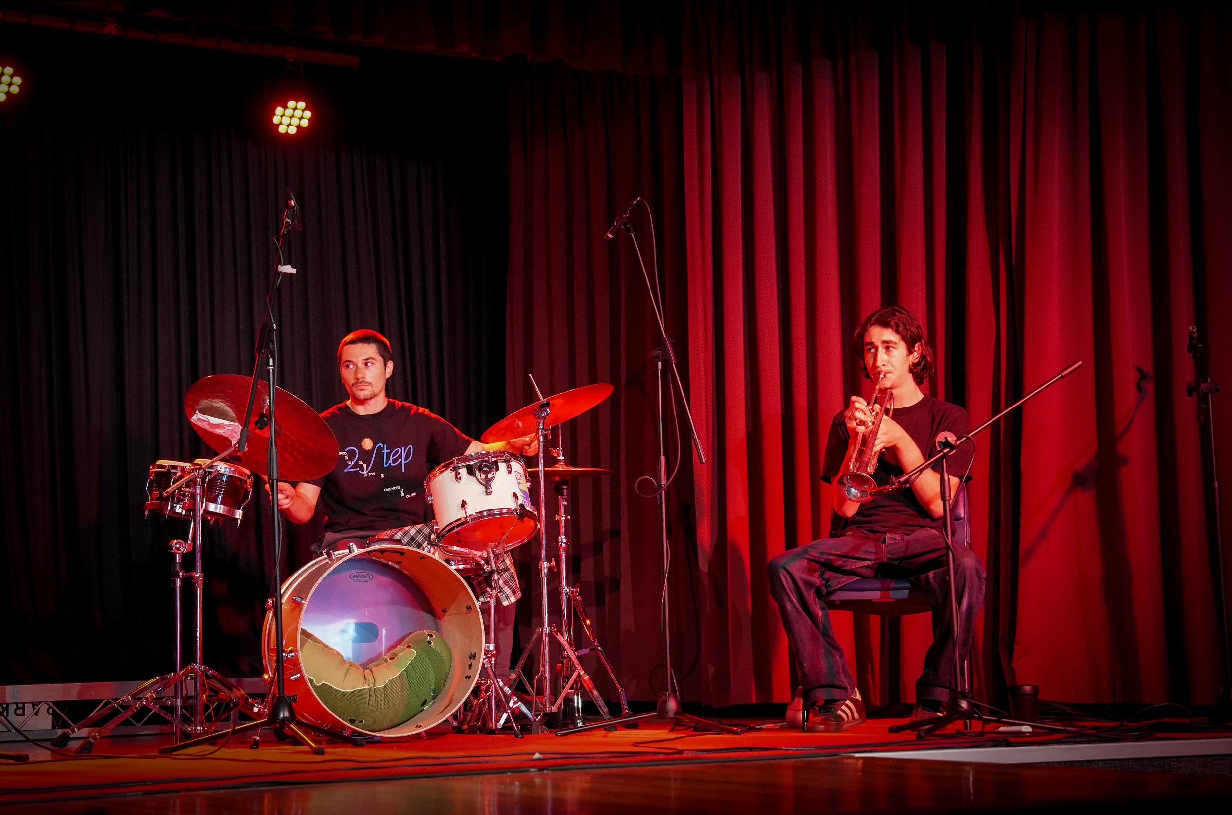 Two musicians on stage, one playing drums and the other playing a trumpet, with red lighting and a dark curtain background.