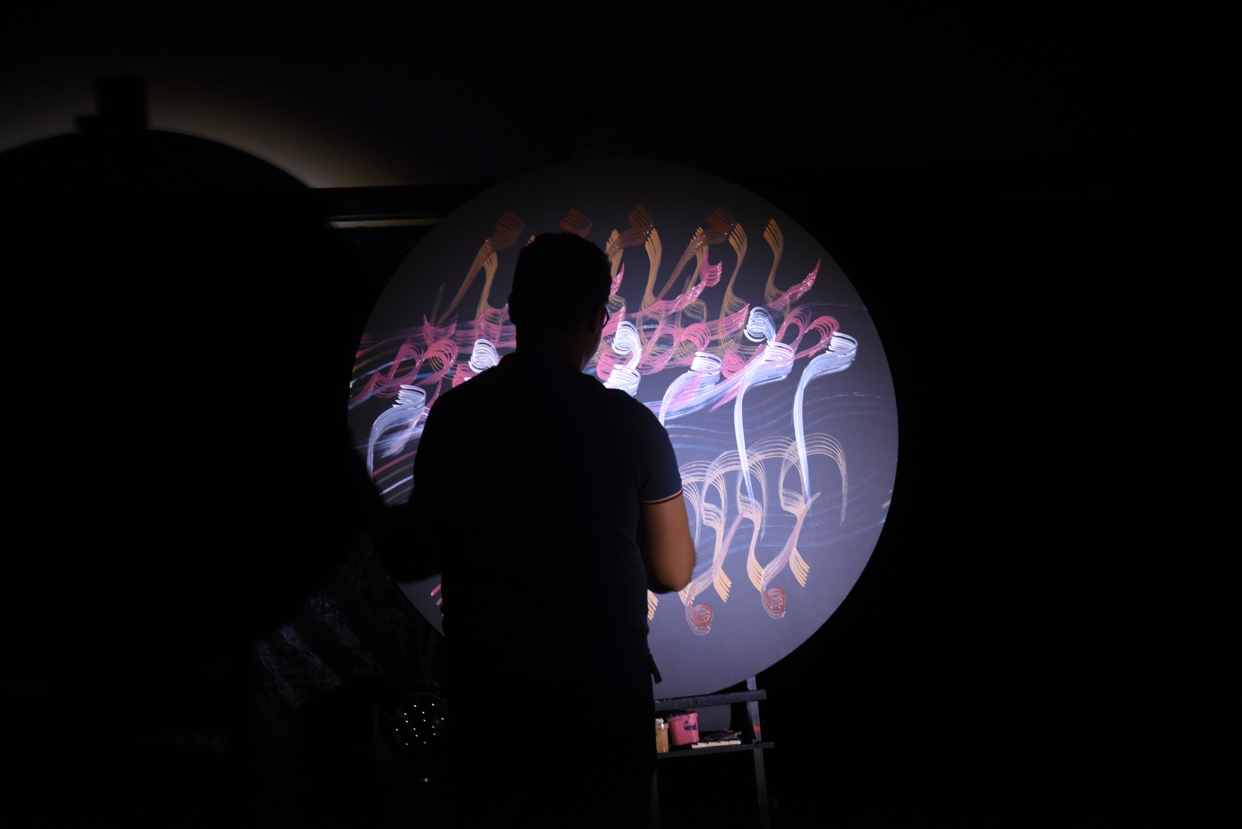 Person painting colorful abstract brushstrokes on a circular canvas in dim lighting.