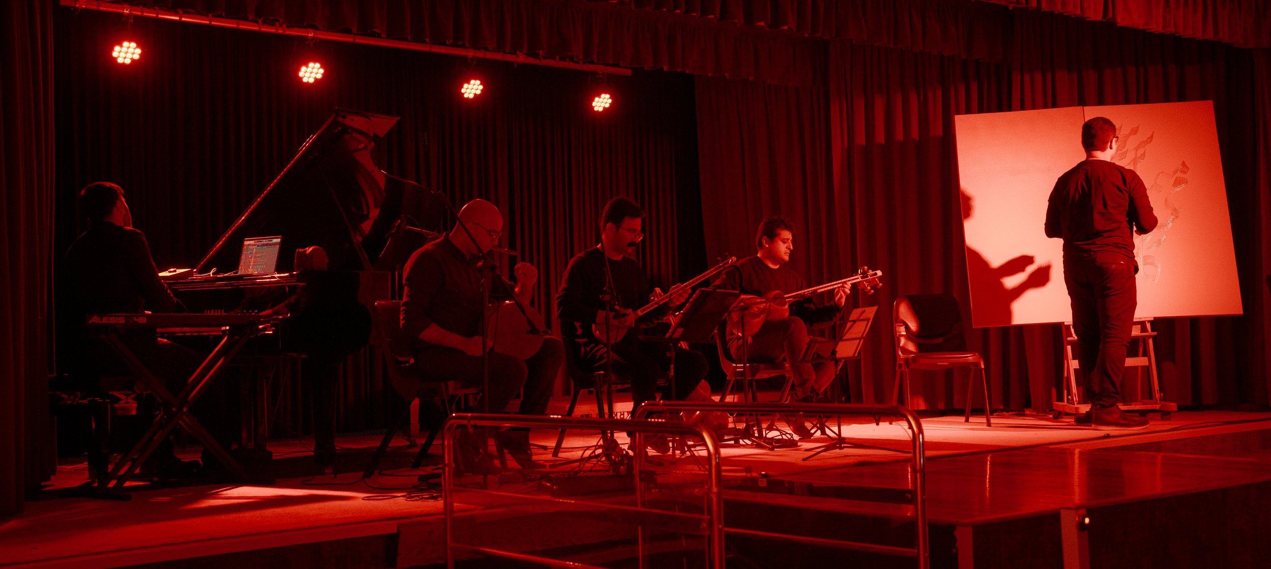 Musicians performing on stage under red lighting, with various instruments including a keyboard and string instruments, and an artist painting on a large canvas.