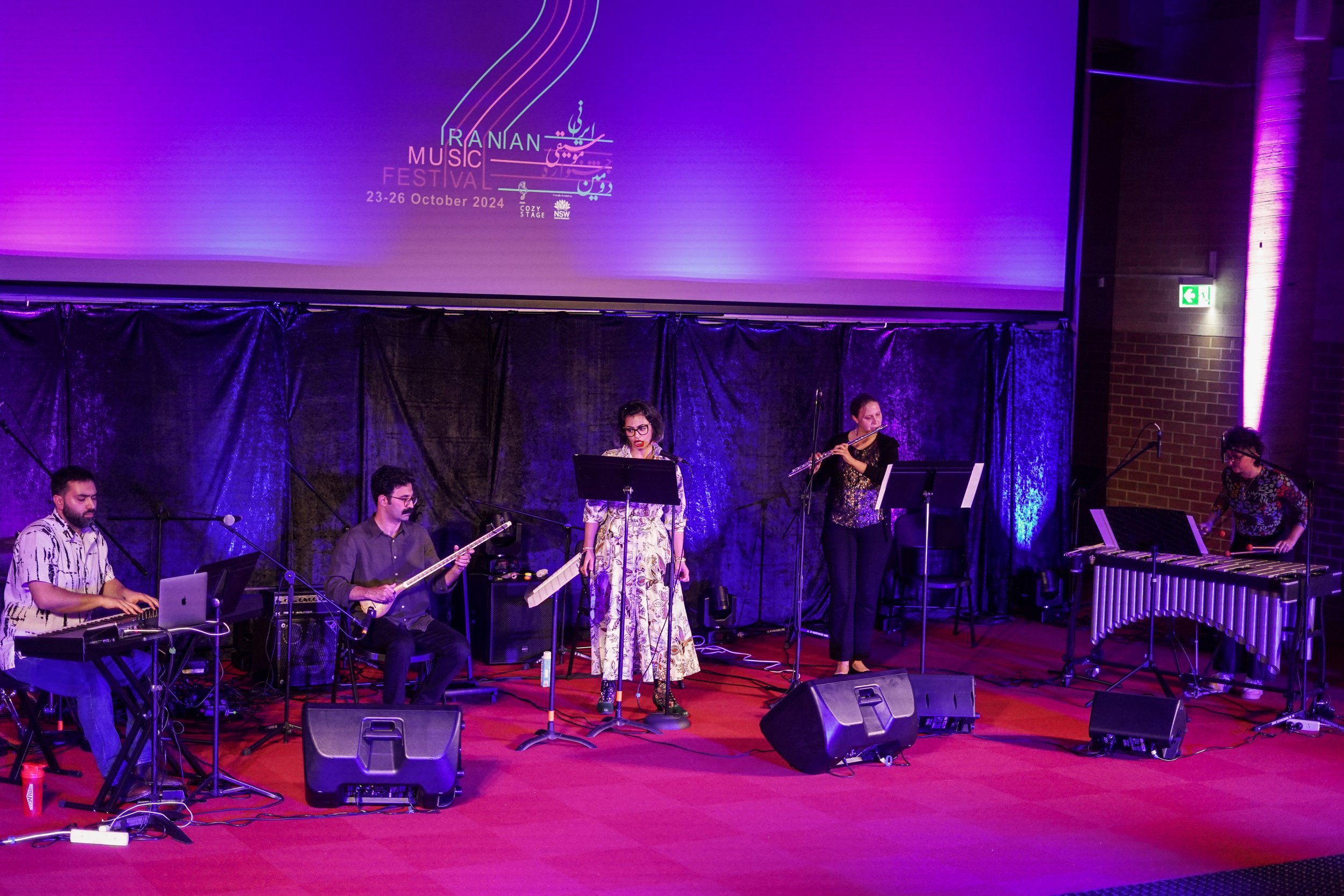 A musical performance on stage with five musicians at the Iranian Music Festival, October 2024. The musicians are playing a keyboard, flute, and other instruments while a singer performs at the center. The stage is lit with purple lights.