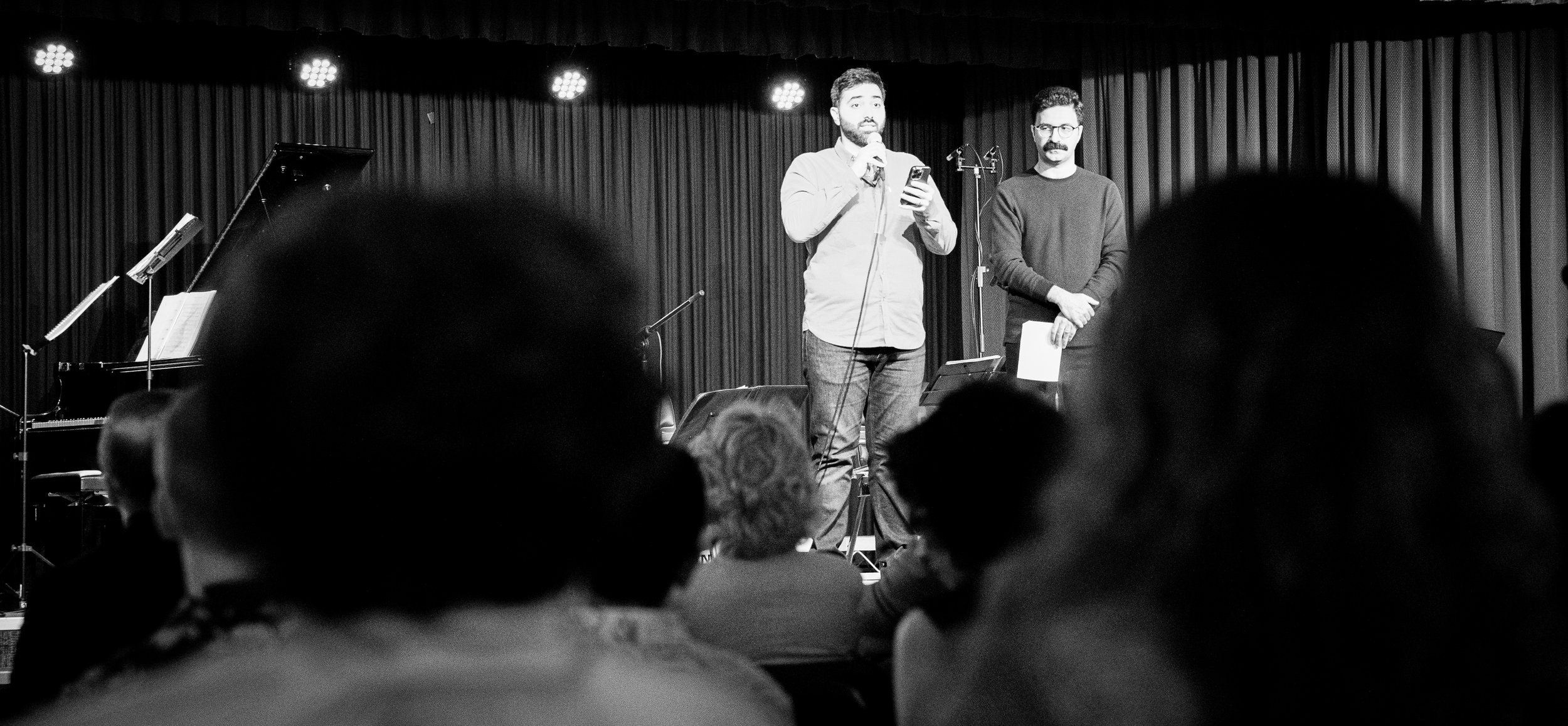 Two men on stage with microphones, addressing an audience; piano visible in background.