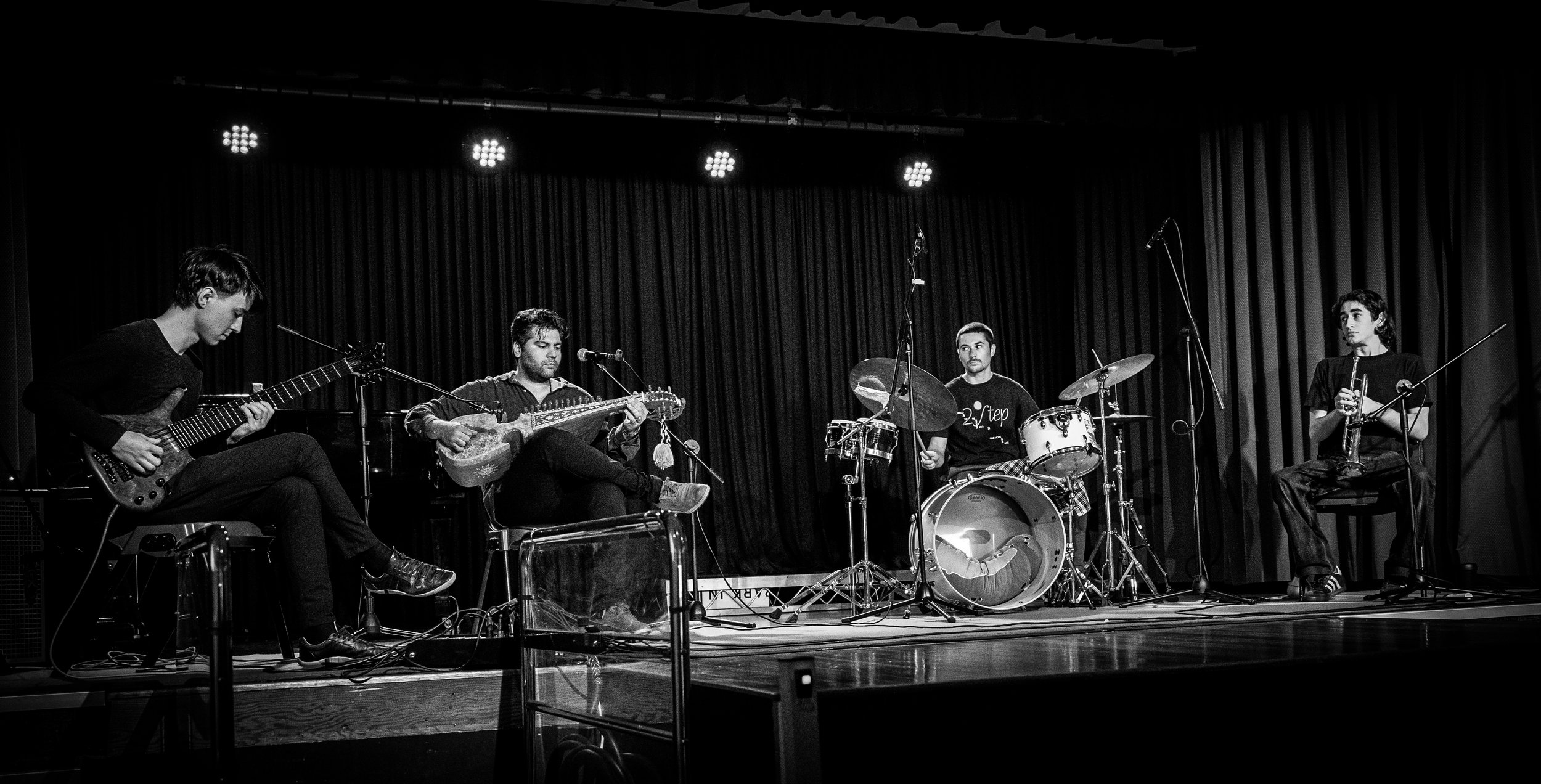 Black and white photo of a band performing on stage with guitar, string instrument, drums, and single-reed woodwind.