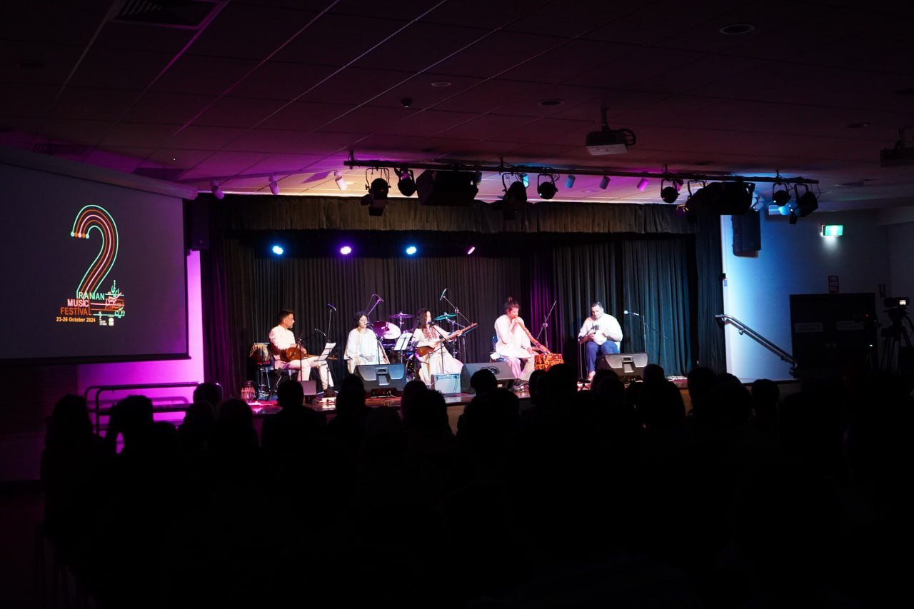 Concert at Iranian Music Festival with musicians on stage, audience in foreground, and event banner on side screen.