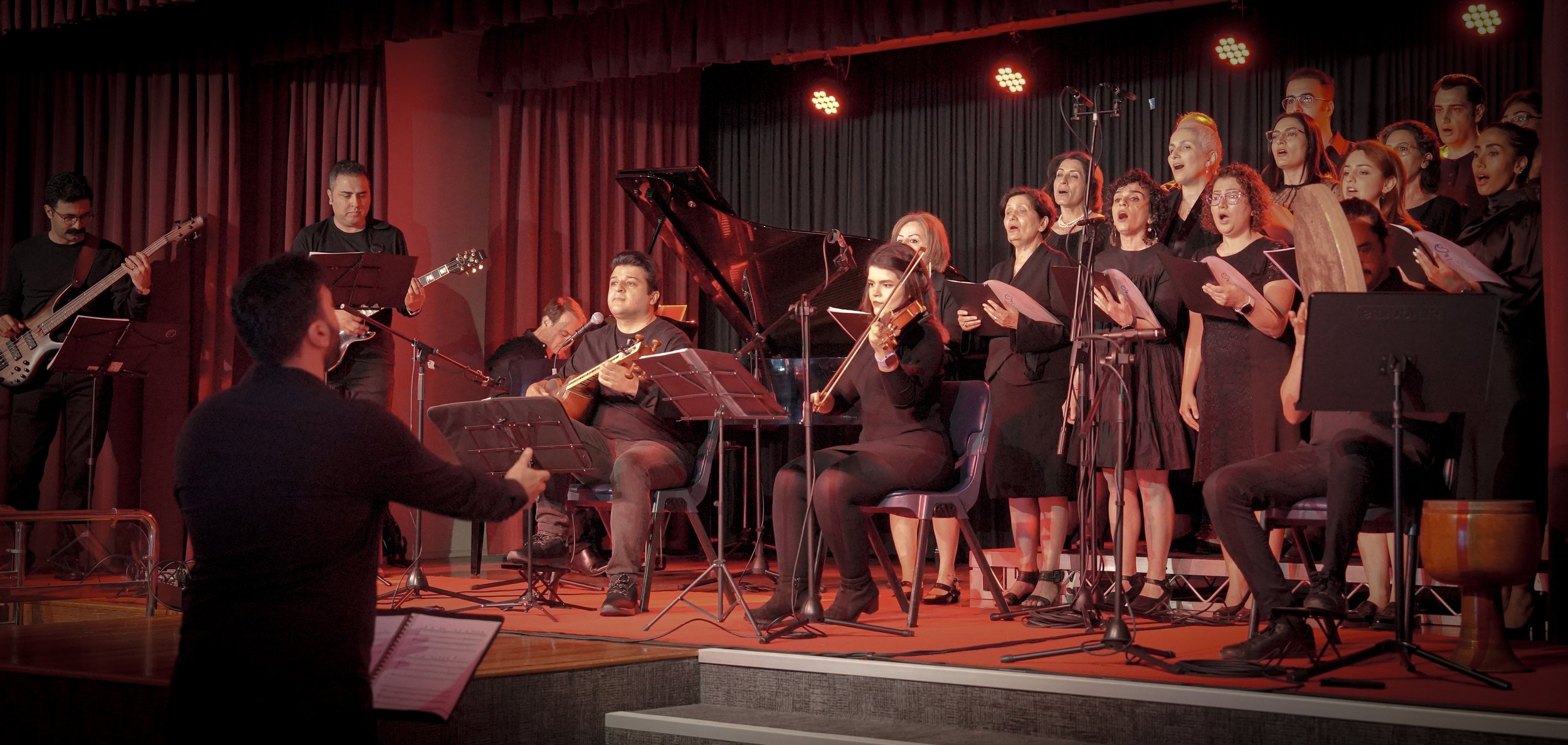 A group of musicians and singers performing on stage with various instruments, including guitar, violin, and piano, and a conductor leading them. The singers are dressed in black and holding sheet music.